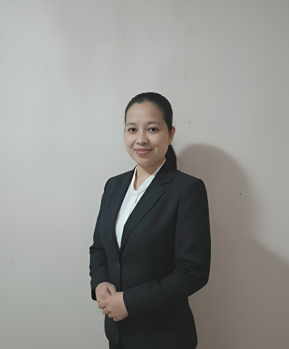 Woman in a black suit smiling, hands clasped, standing against a plain light wall.