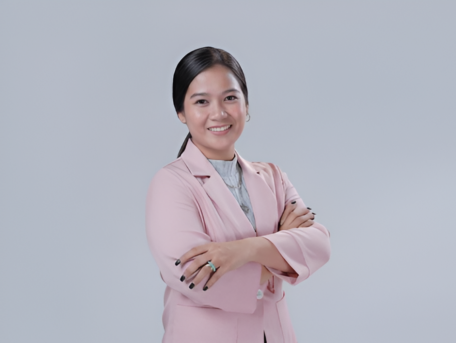 Woman in pink blazer smiles, arms crossed, gray background.