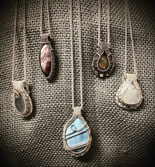 Five teardrop pendant necklaces on textured fabric, with stones in blue, white, amber, and mauve.