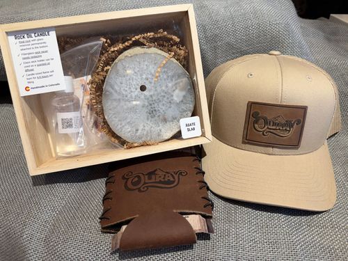 Gift box with hat, tumbler, wood slice, and leather wallet beside a tan cap on a gray surface