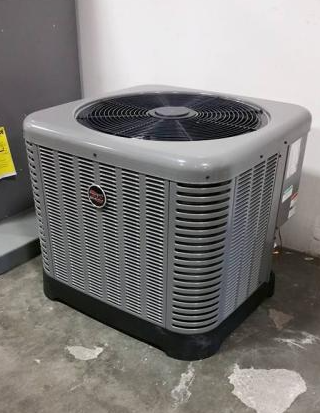 A grey Rheem outdoor air conditioning unit sits on a concrete floor next to a wall.