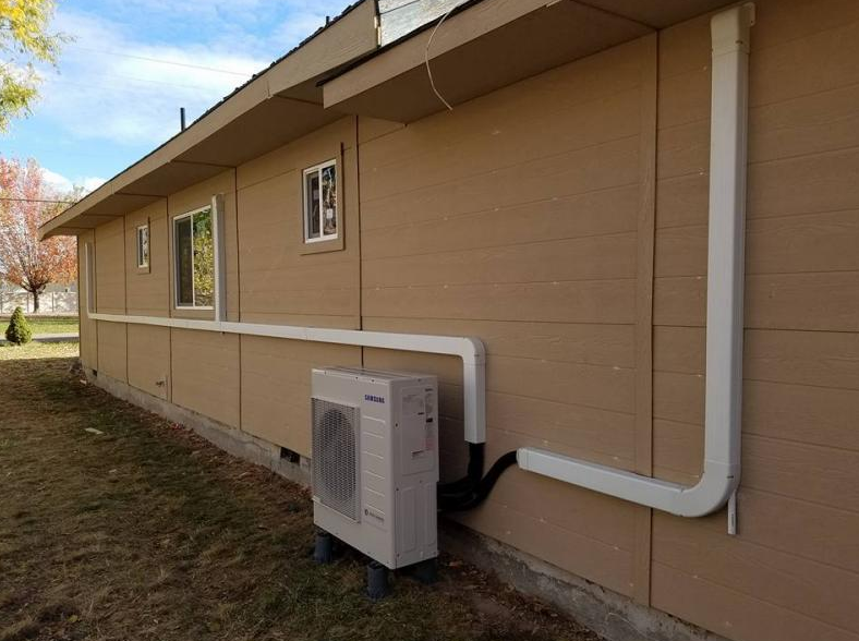 Exterior of a house with a beige wall featuring a grey HVAC condenser unit connected to white conduit pipes.