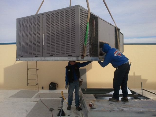 Two workers guide a large HVAC unit suspended by a crane onto a rooftop.