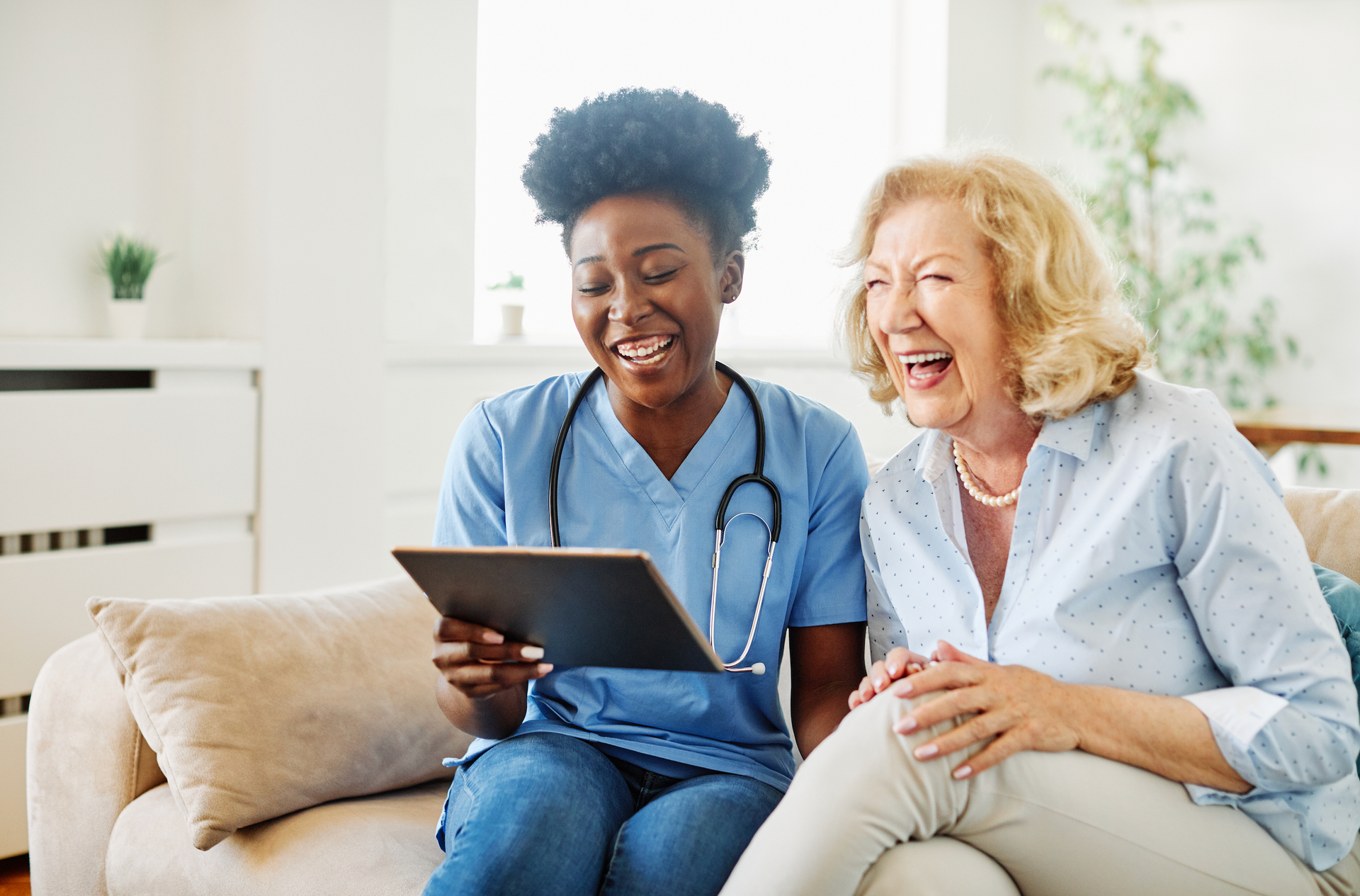 A nurse and an elderly woman are sitting on a couch looking at a tablet.