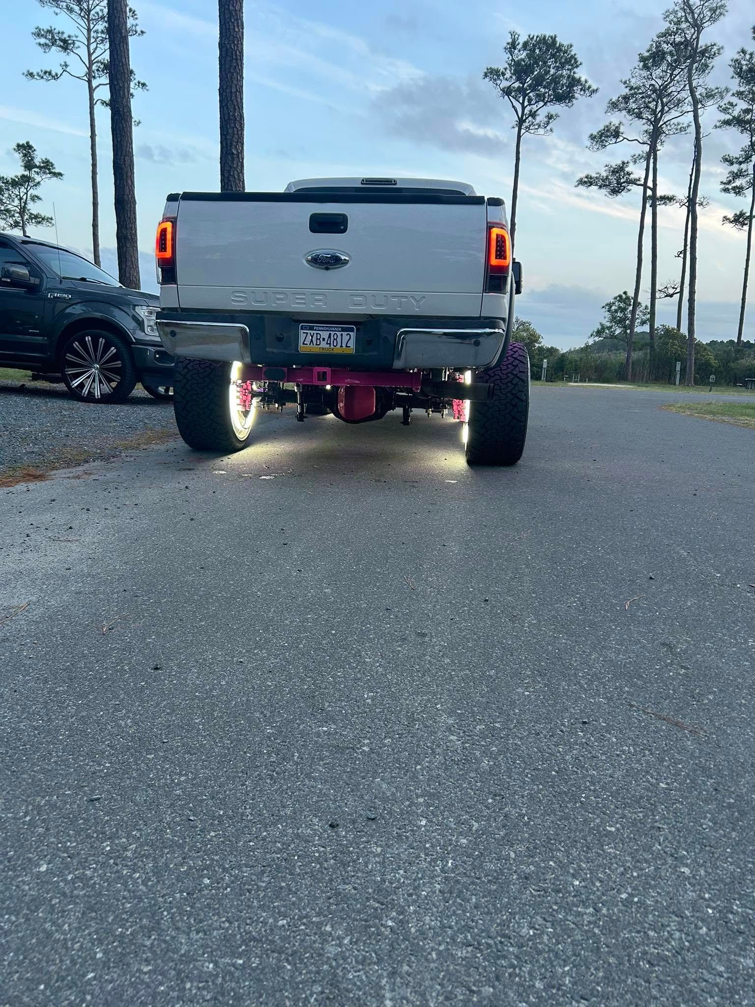 White truck with illuminated pink undercarriage on an asphalt road. Black vehicle is parked on the left.