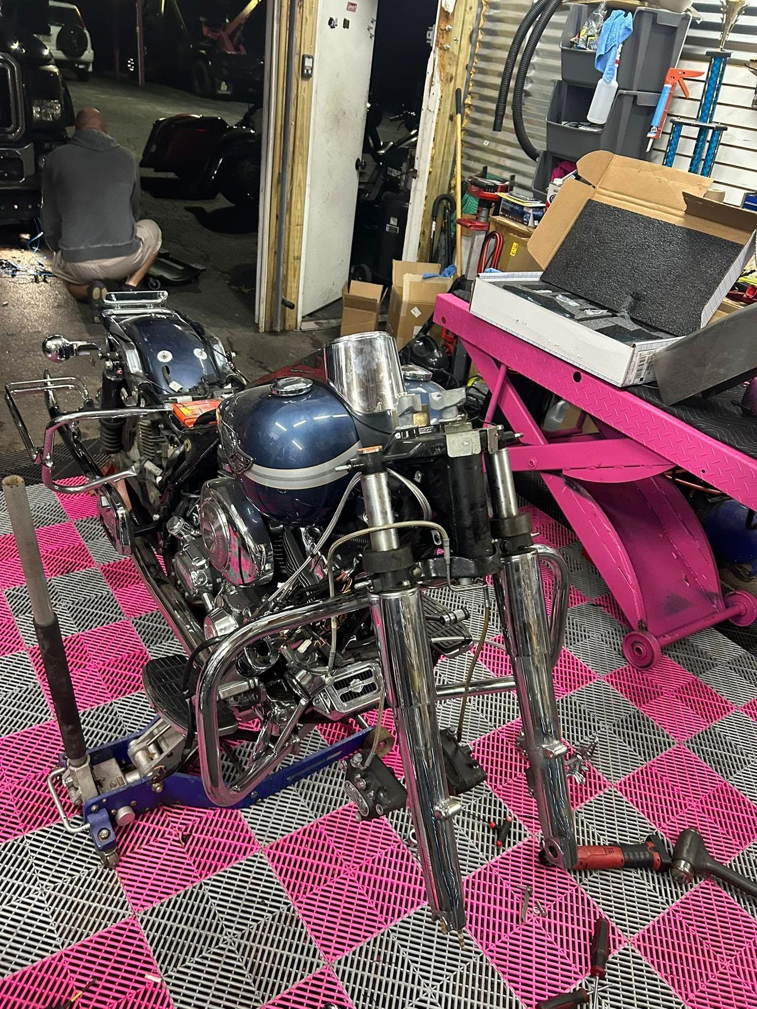 Motorcycle being worked on in a garage; a person kneels nearby, surrounded by tools and equipment.