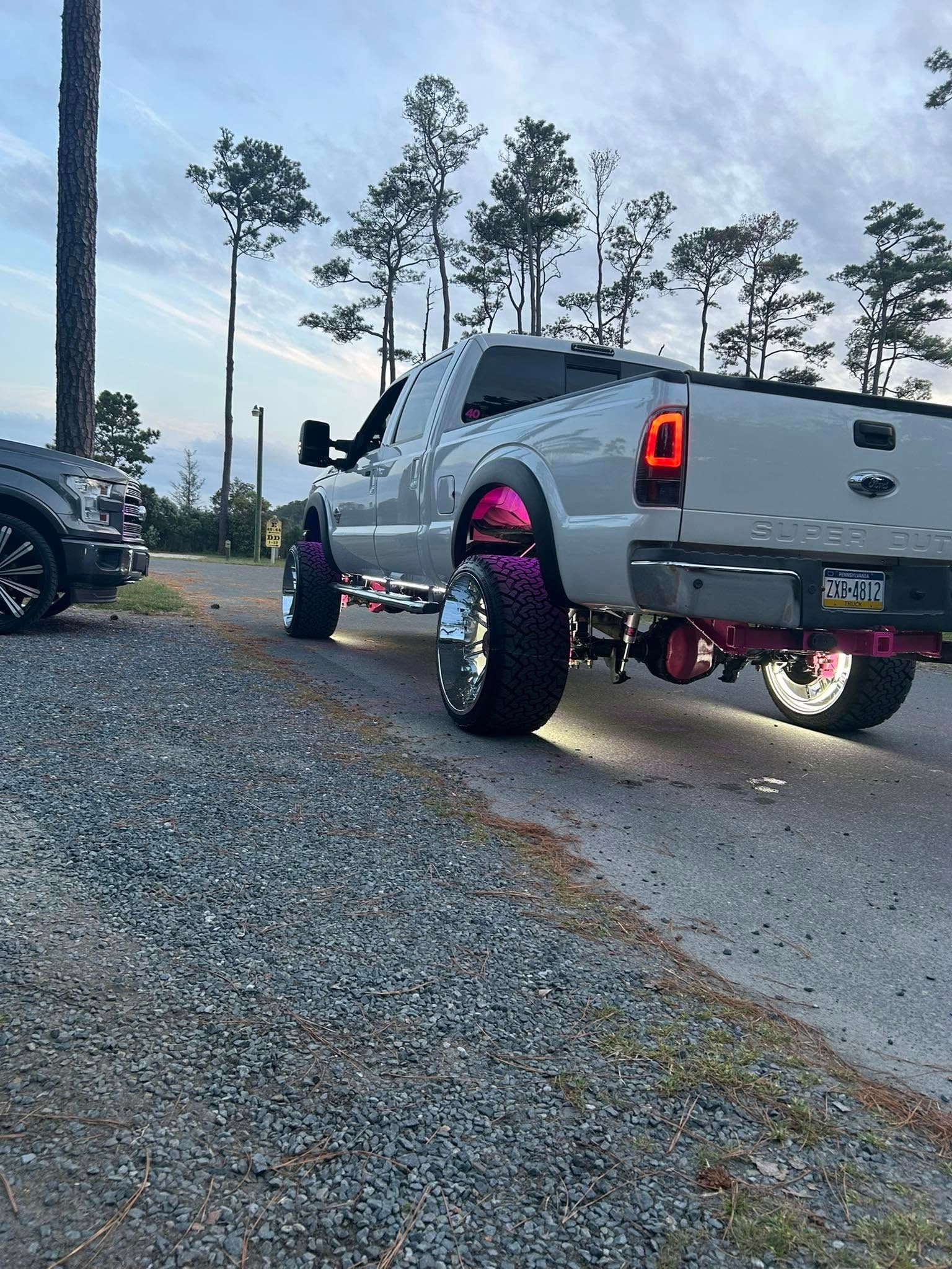 White lifted truck with pink accents, big wheels, parked on gravel road.