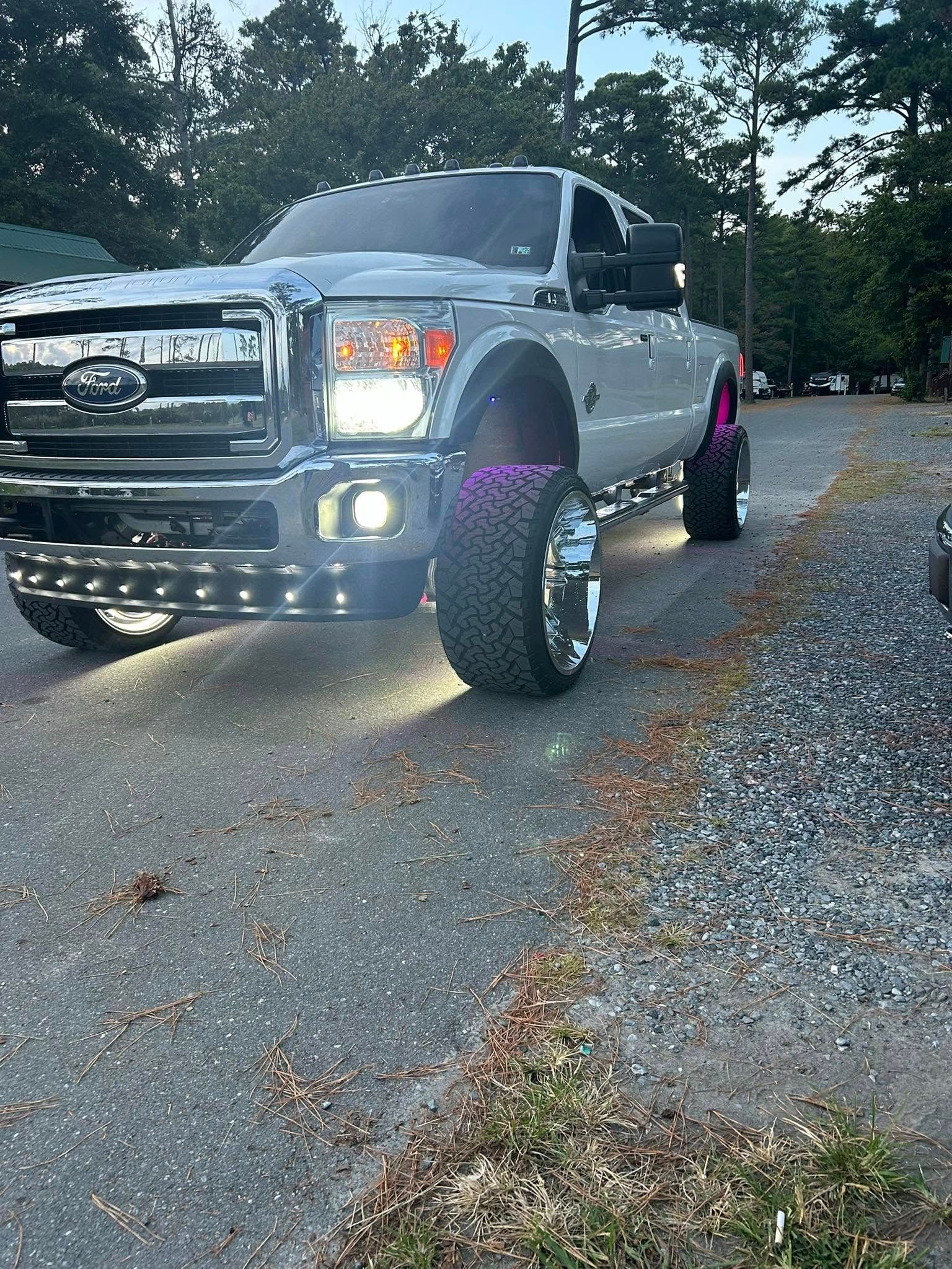 White Ford truck with chrome detailing, large tires, and LED lights, parked on a gravel road.