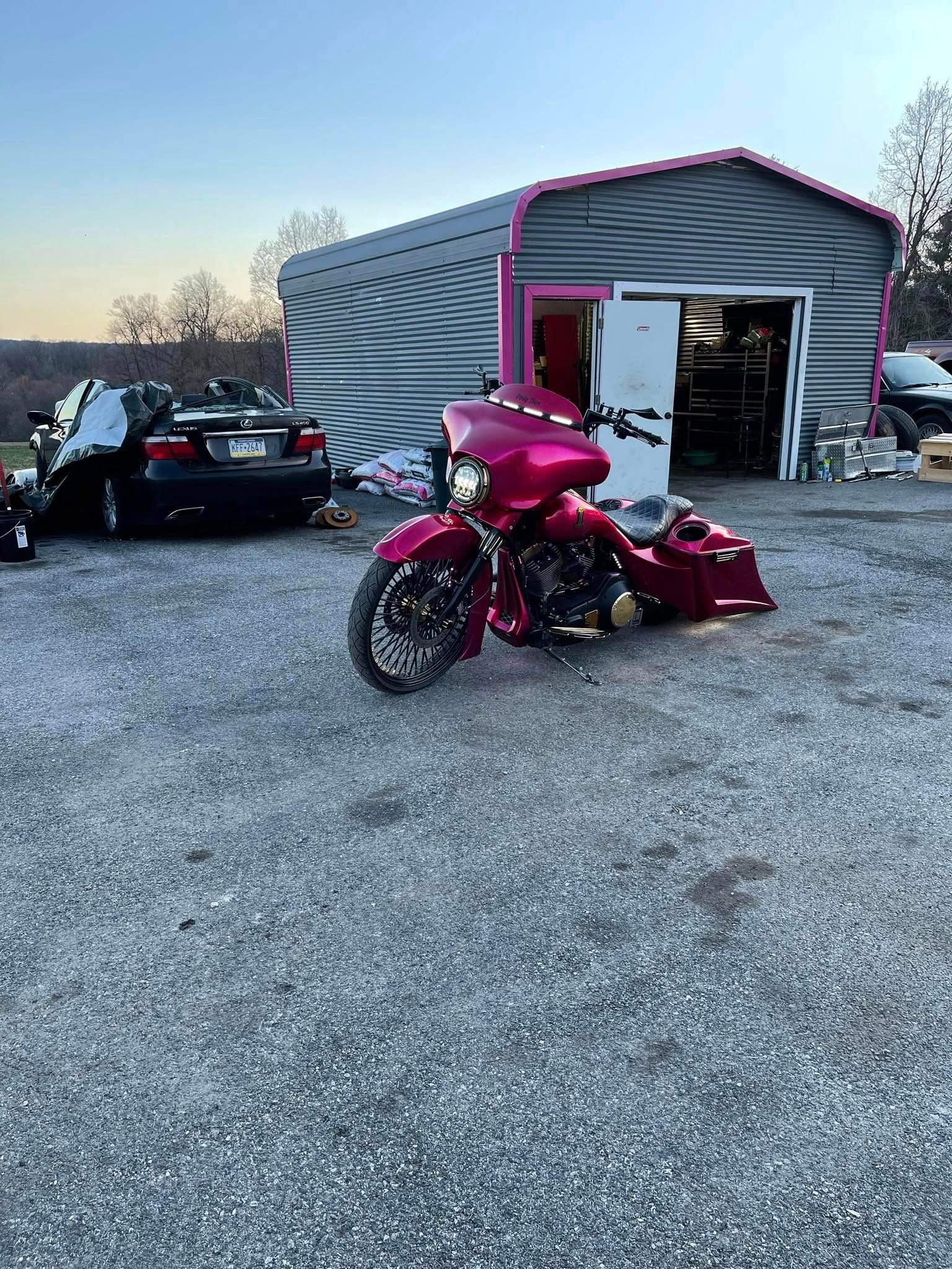 Pink motorcycle parked in front of a gray shed with pink trim; black car in the background.