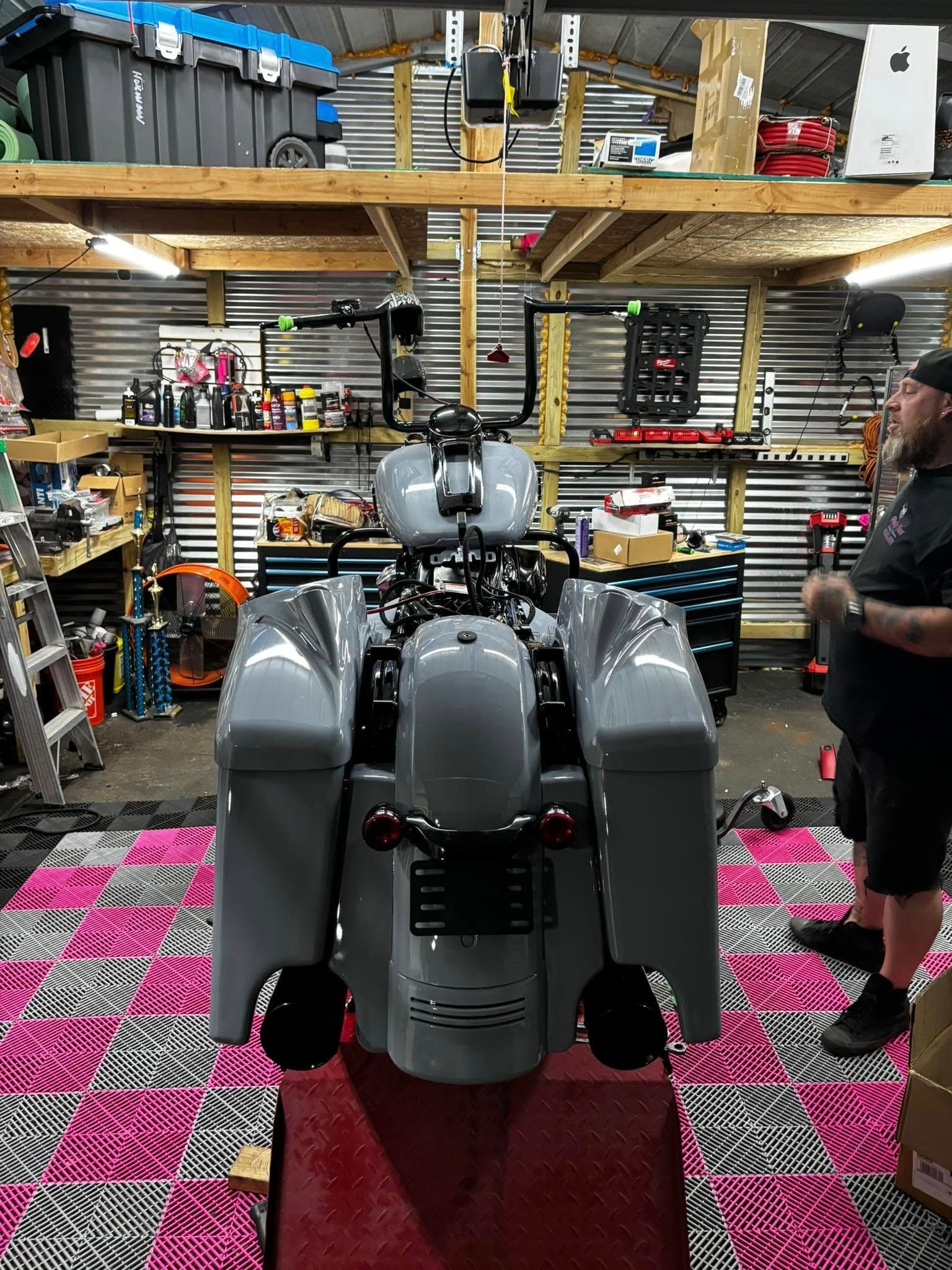 Motorcycle in a garage, gray paint, custom handlebars, man standing nearby, tool storage in background.