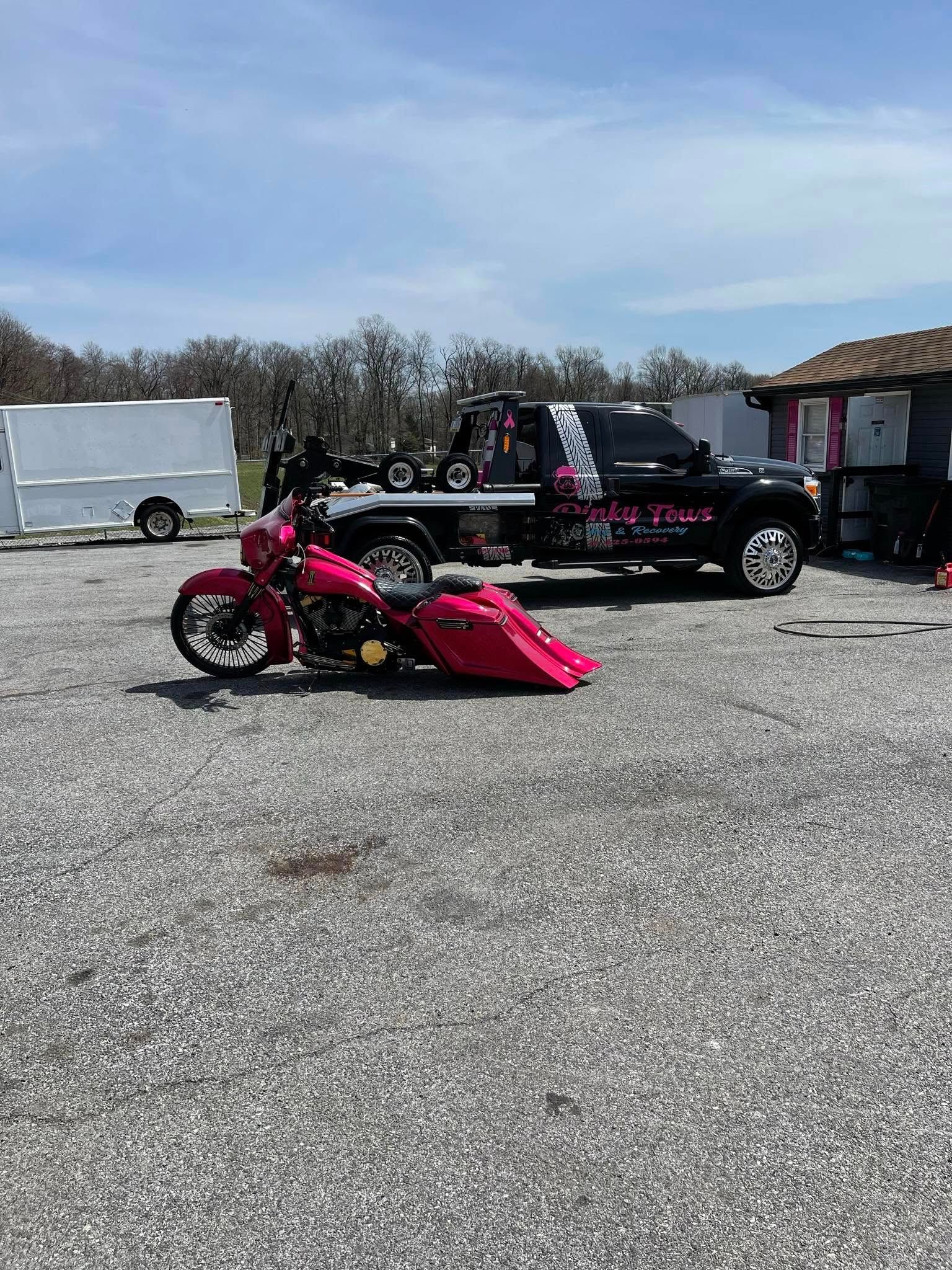 Pink motorcycle with a detached fuel tank next to a tow truck in a gravel lot.