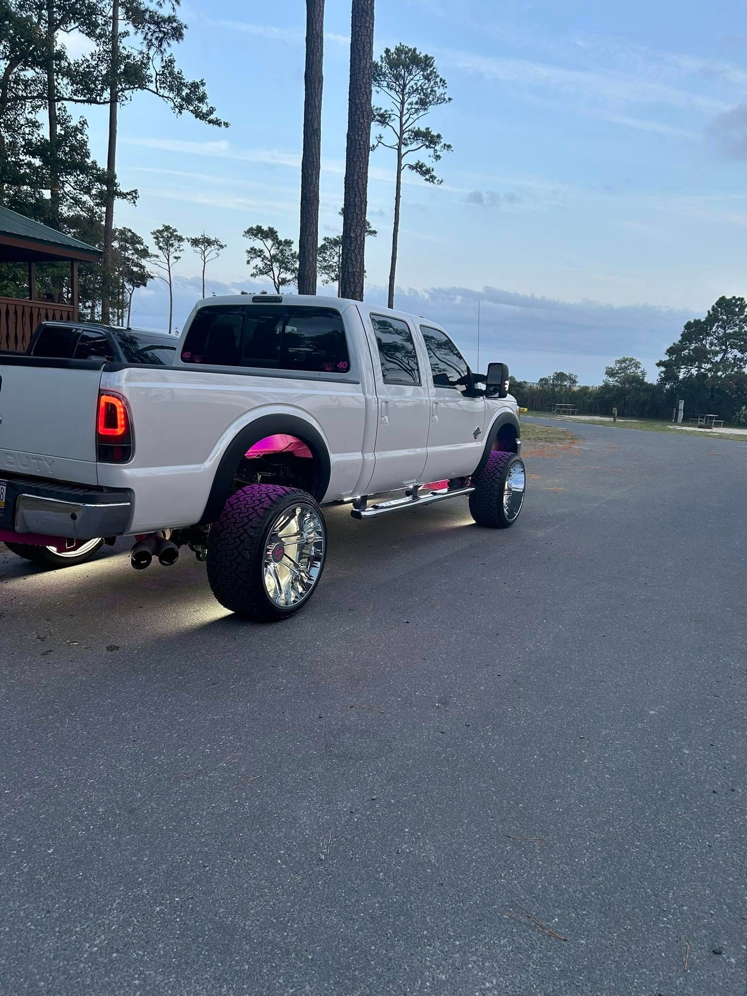 White truck with chrome wheels and pink accents parked on asphalt. Trees in the background.