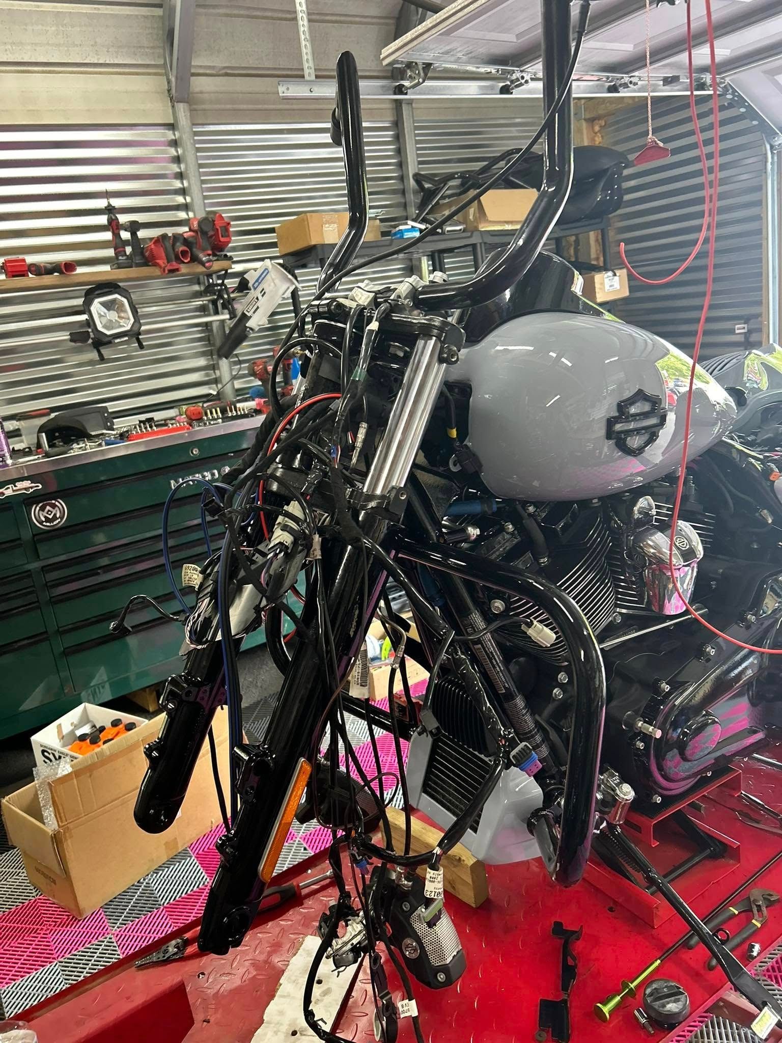 Black motorcycle in a garage, handlebars detached, wires exposed, tools and parts visible.