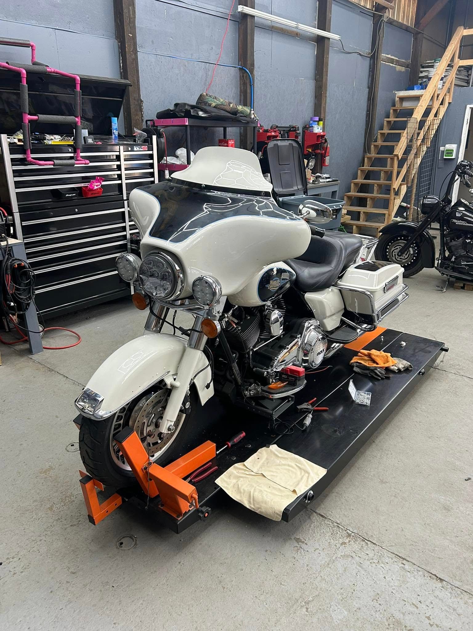 White and black motorcycle on a lift in a workshop. Tools and stairs are visible in the background.
