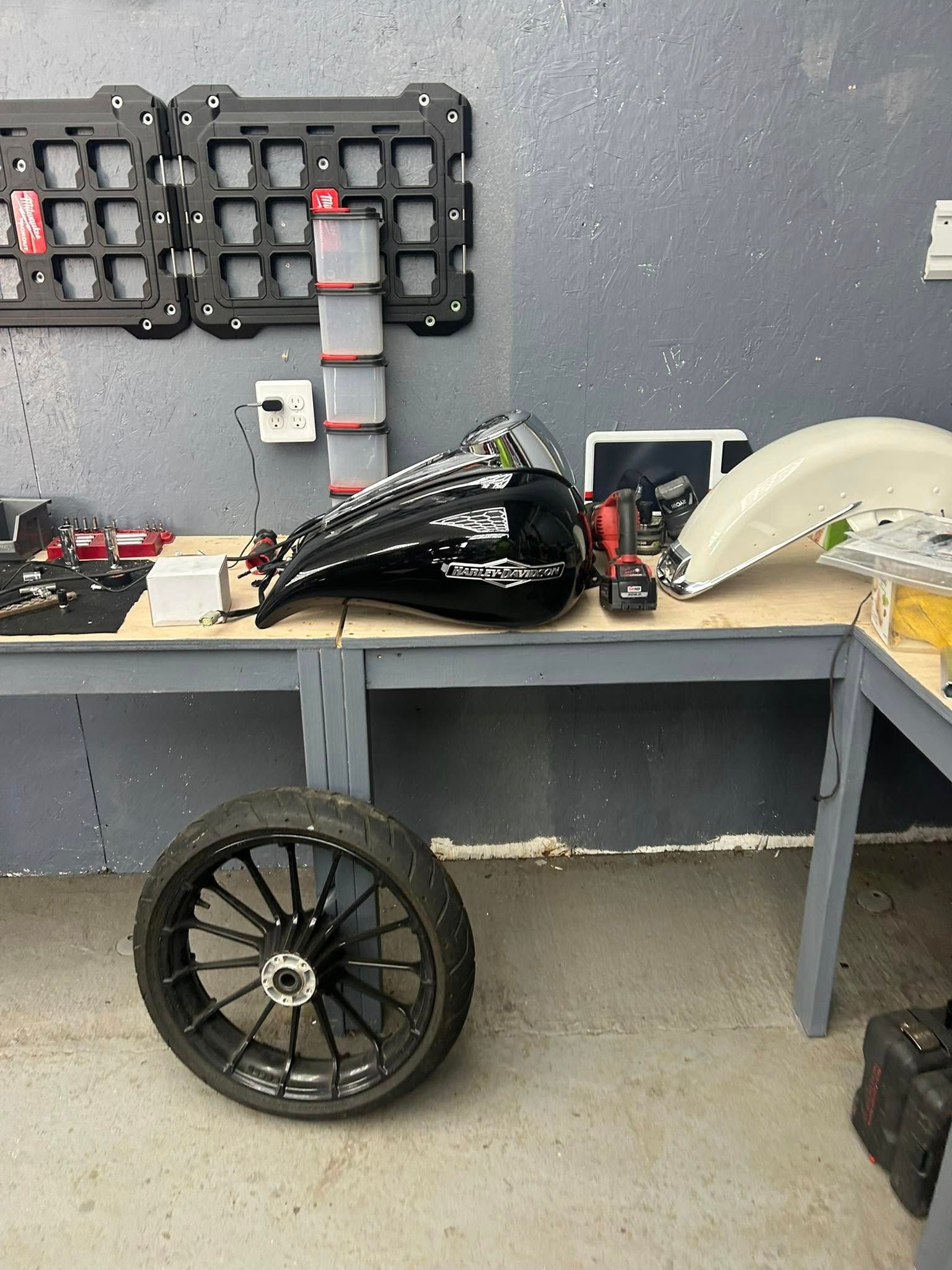 Motorcycle parts, including a gas tank, wheel, and fender, are on a workbench in a garage.