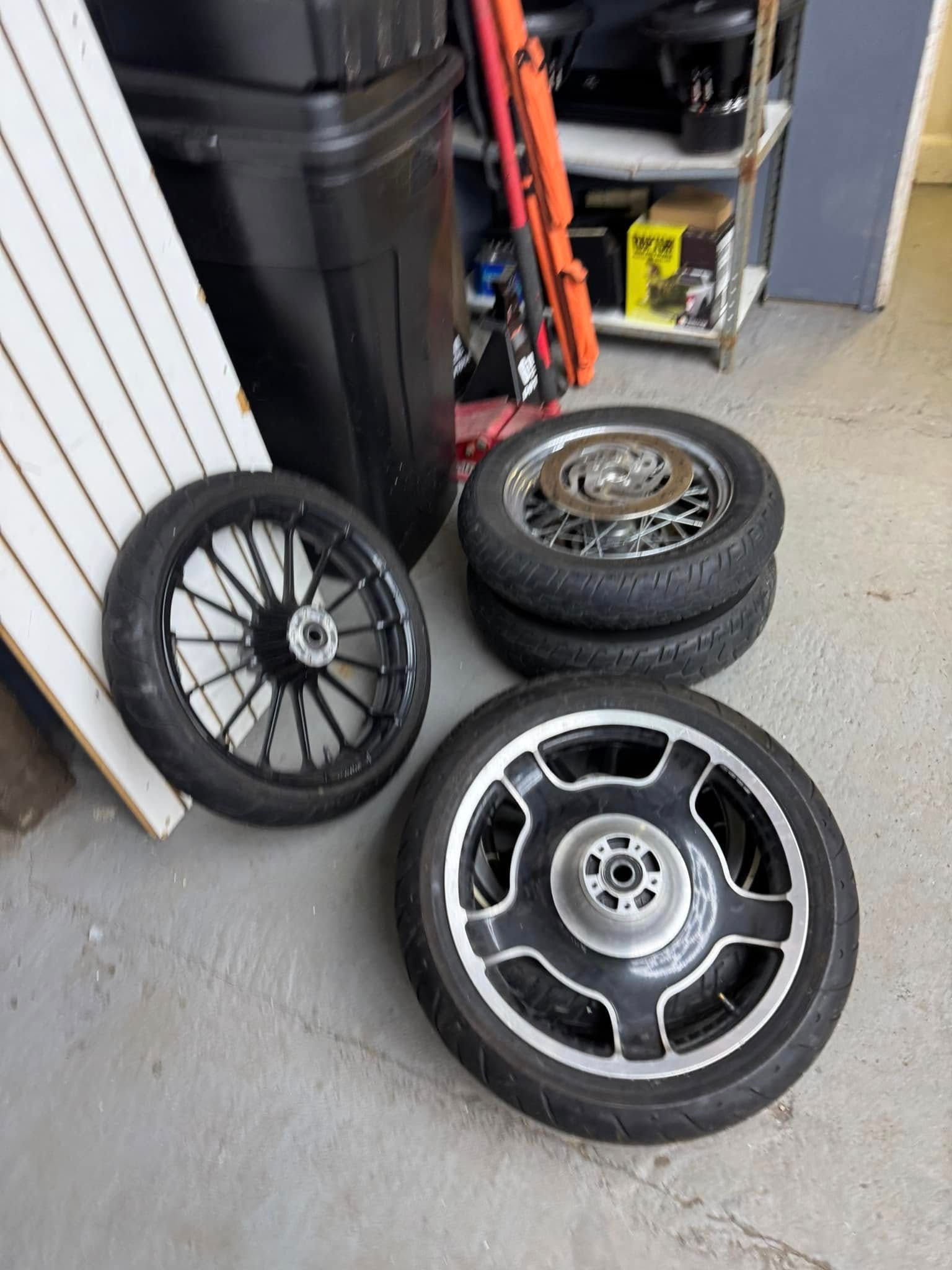 Four motorcycle wheels on a concrete floor next to a white panel and storage bins.