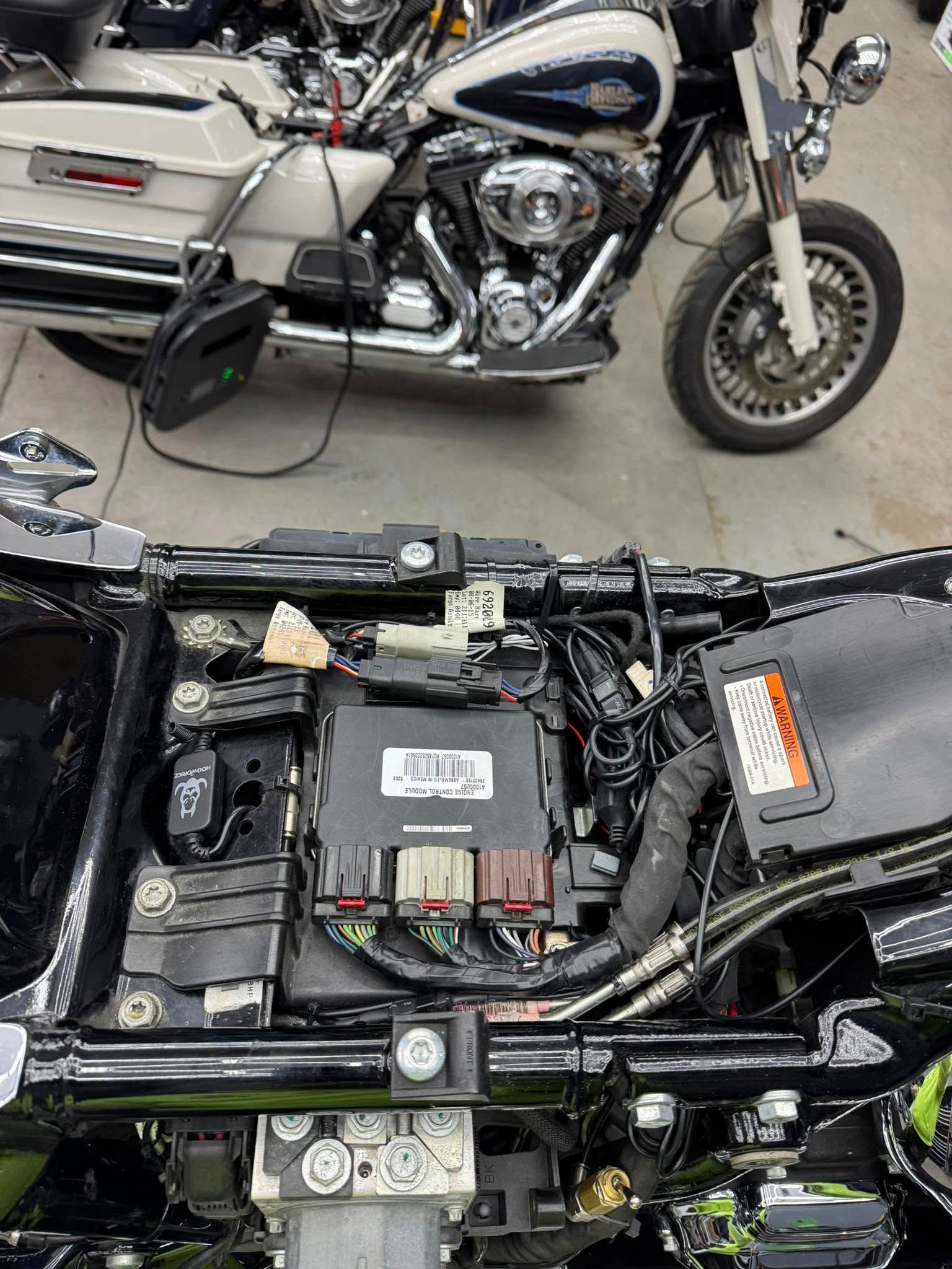 Motorcycle's wiring exposed with a second motorcycle in the background. Black, silver, and cream colors.