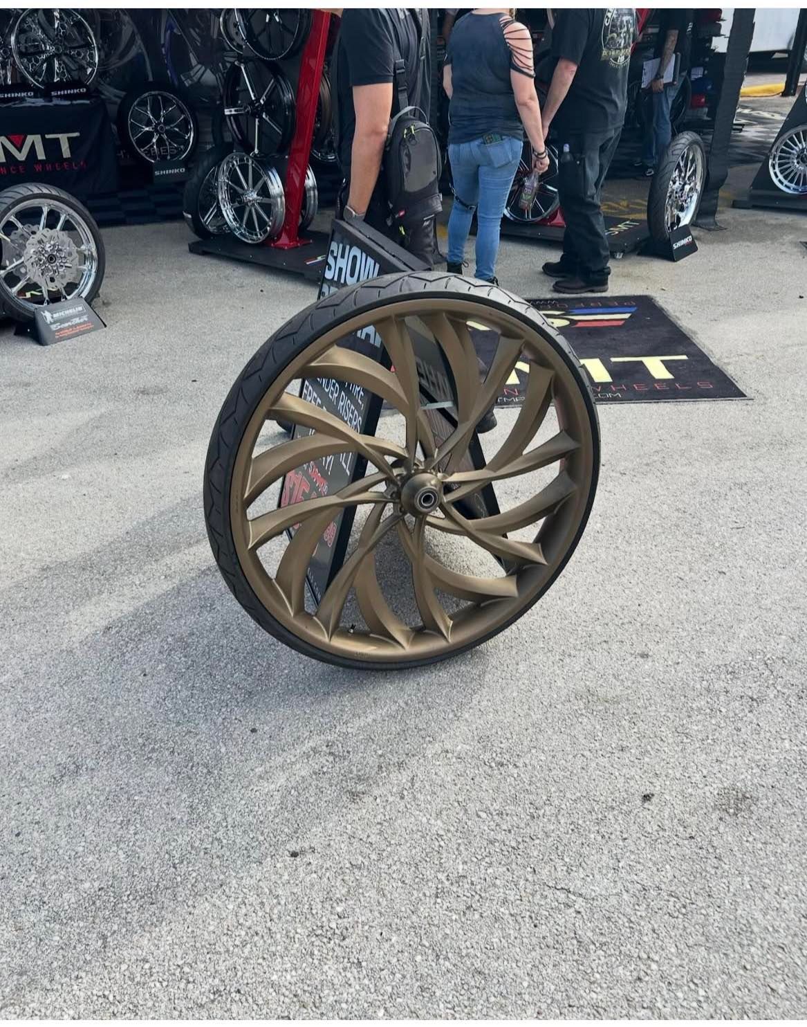Bronze-colored custom car wheel on display at an outdoor event, people and other wheels visible in the background.