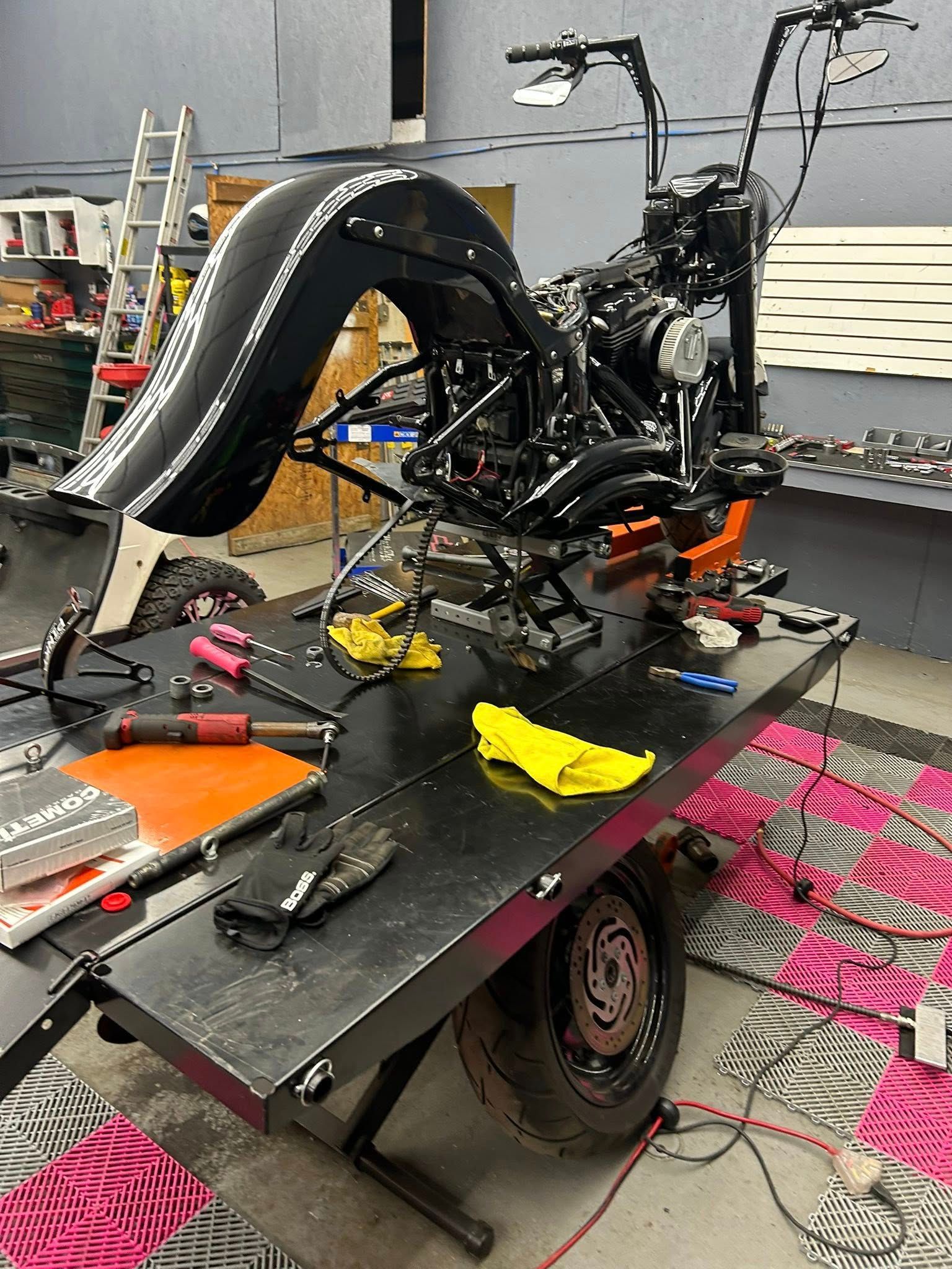 Black motorcycle in progress on a black lift table, surrounded by tools and a pink patterned rug.