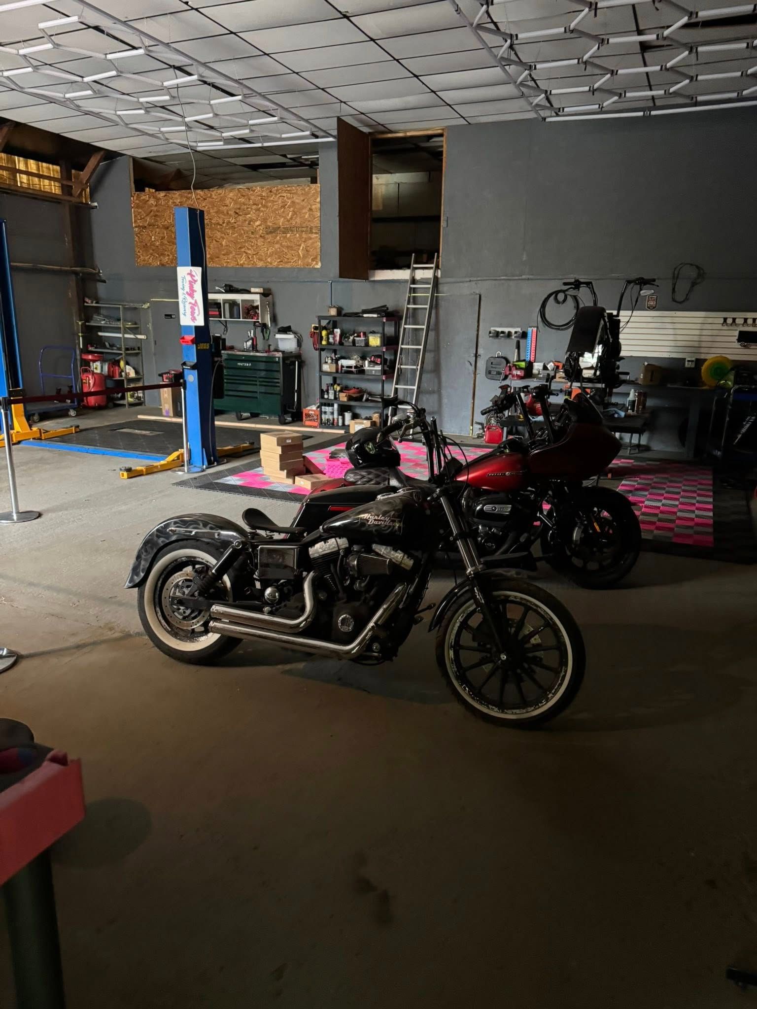 Motorcycles inside a garage, one black, one red. The garage has tools and a lift.