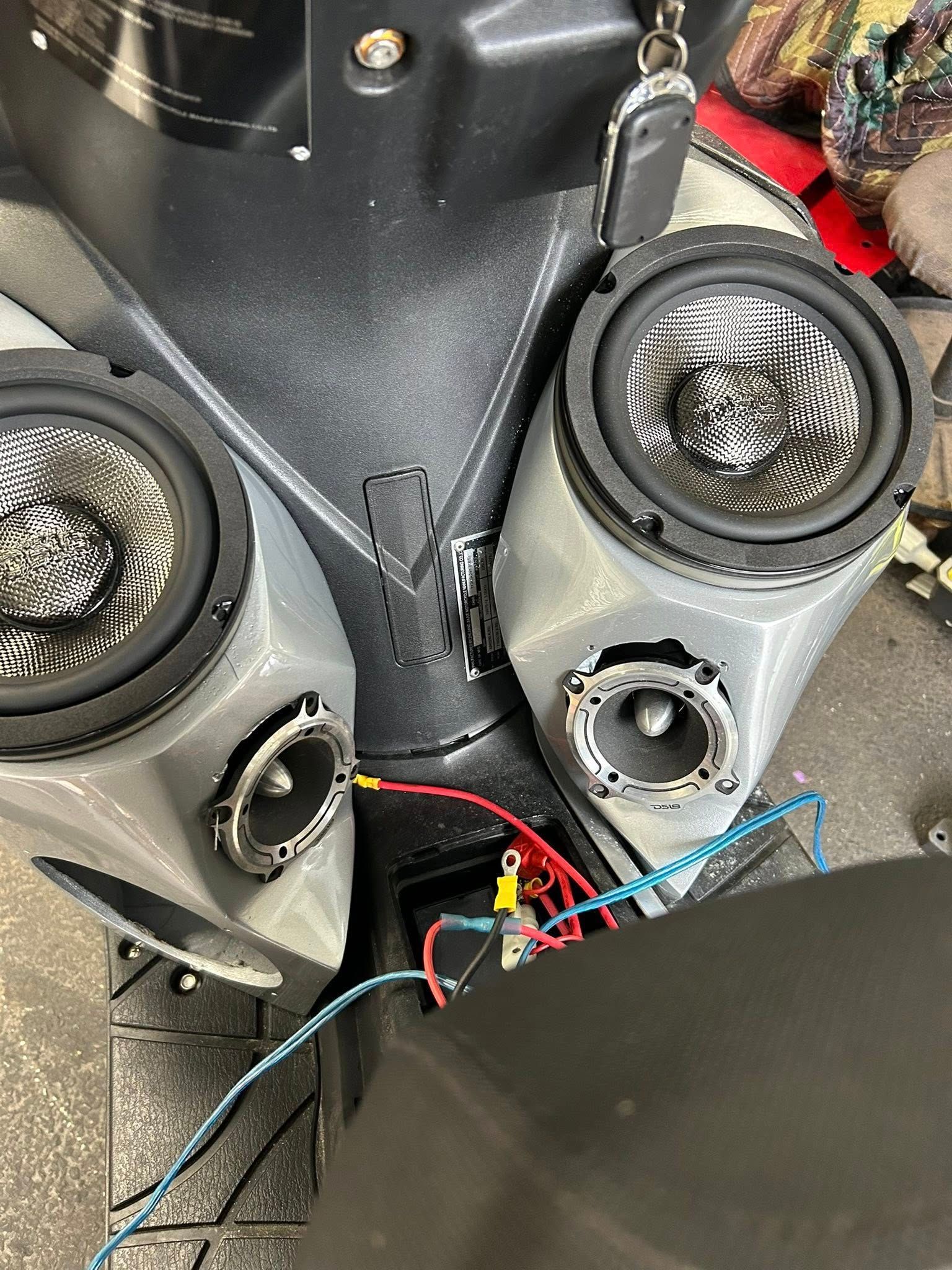Scooter with two speakers installed. Wires connect to a battery. Gray speaker housing with black speakers.