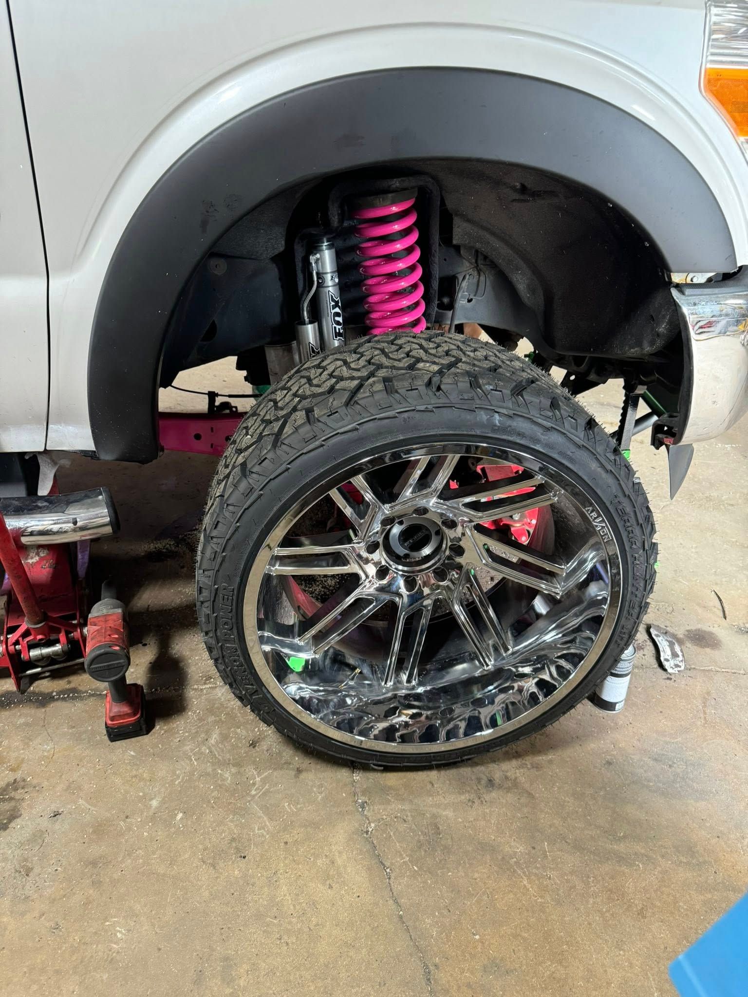 Silver truck wheel with chrome rims and large tire. Pink suspension and black fender.