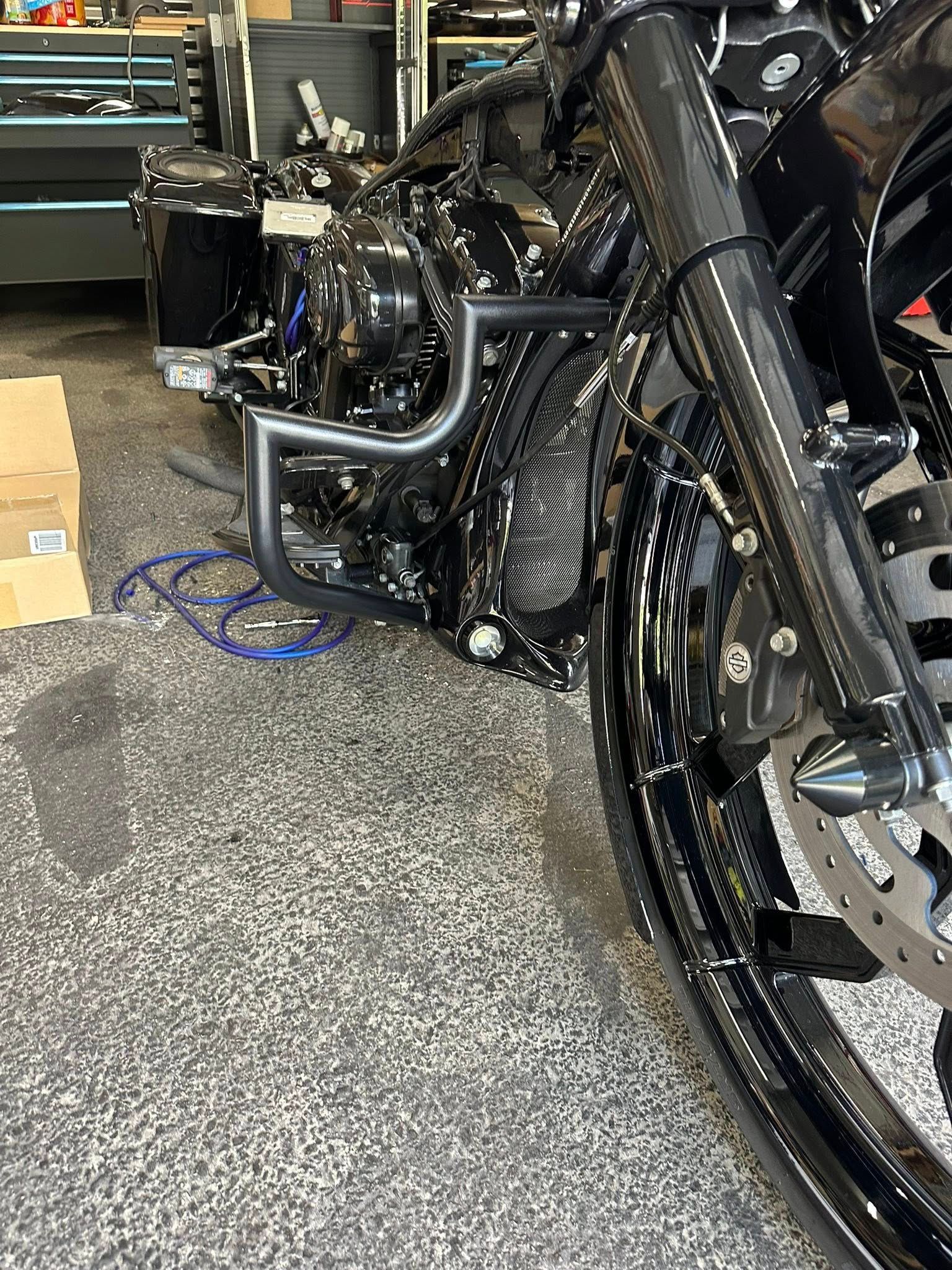 Black motorcycle front section, shiny finish. Parts visible, including forks, wheel, and frame. Inside a shop.