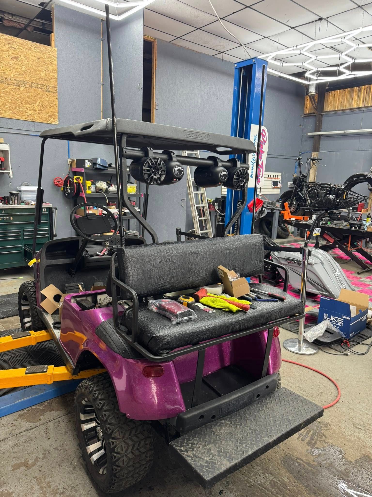 Purple golf cart in a garage, modified with speakers, lifted on a ramp. Tools and equipment visible.