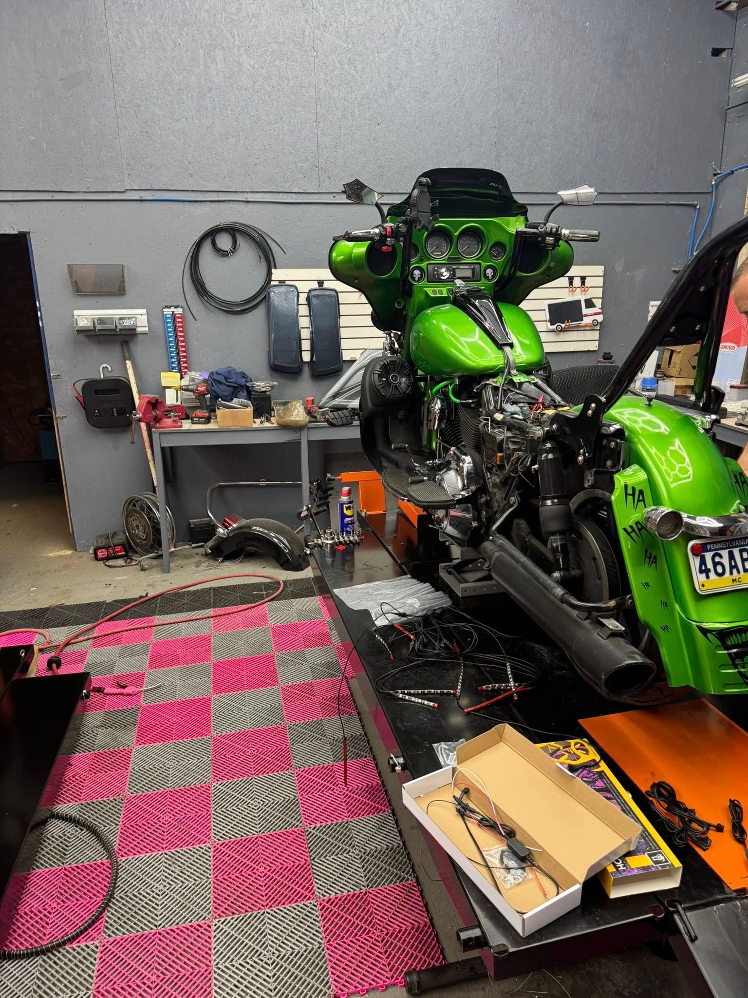 Green motorcycle being worked on in a garage; parts and tools scattered on a pink and black checkered floor.
