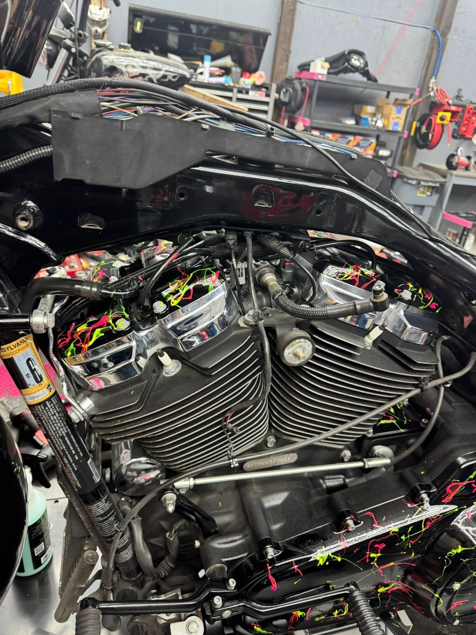Motorcycle engine with chrome and black details, covered in colorful paint splatters. Workshop setting.