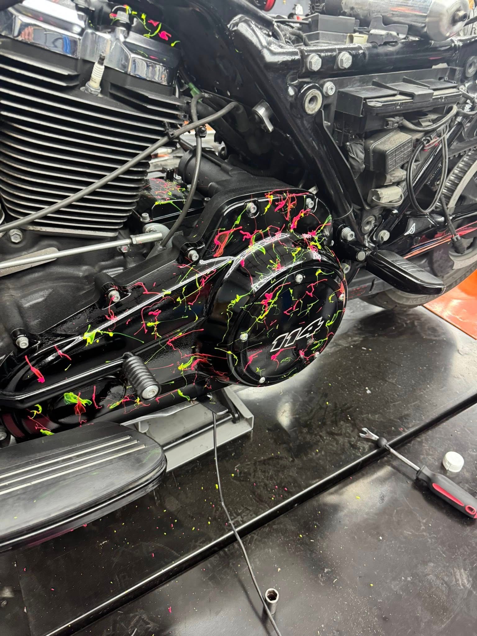 Motorcycle engine with black cover, multi-colored paint splatters, and a screwdriver on a workbench.