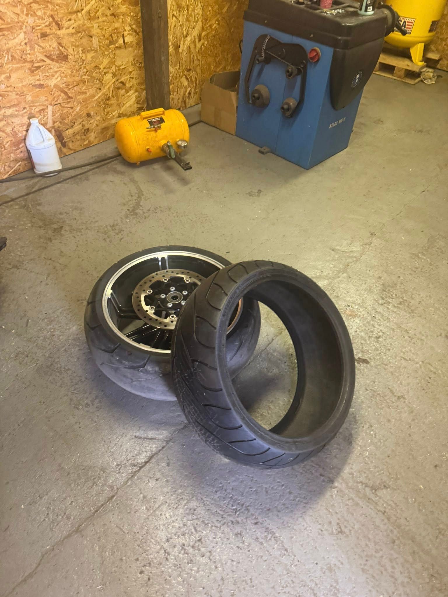 Motorcycle tire and wheel on garage floor; balancing machine and other equipment in the background.