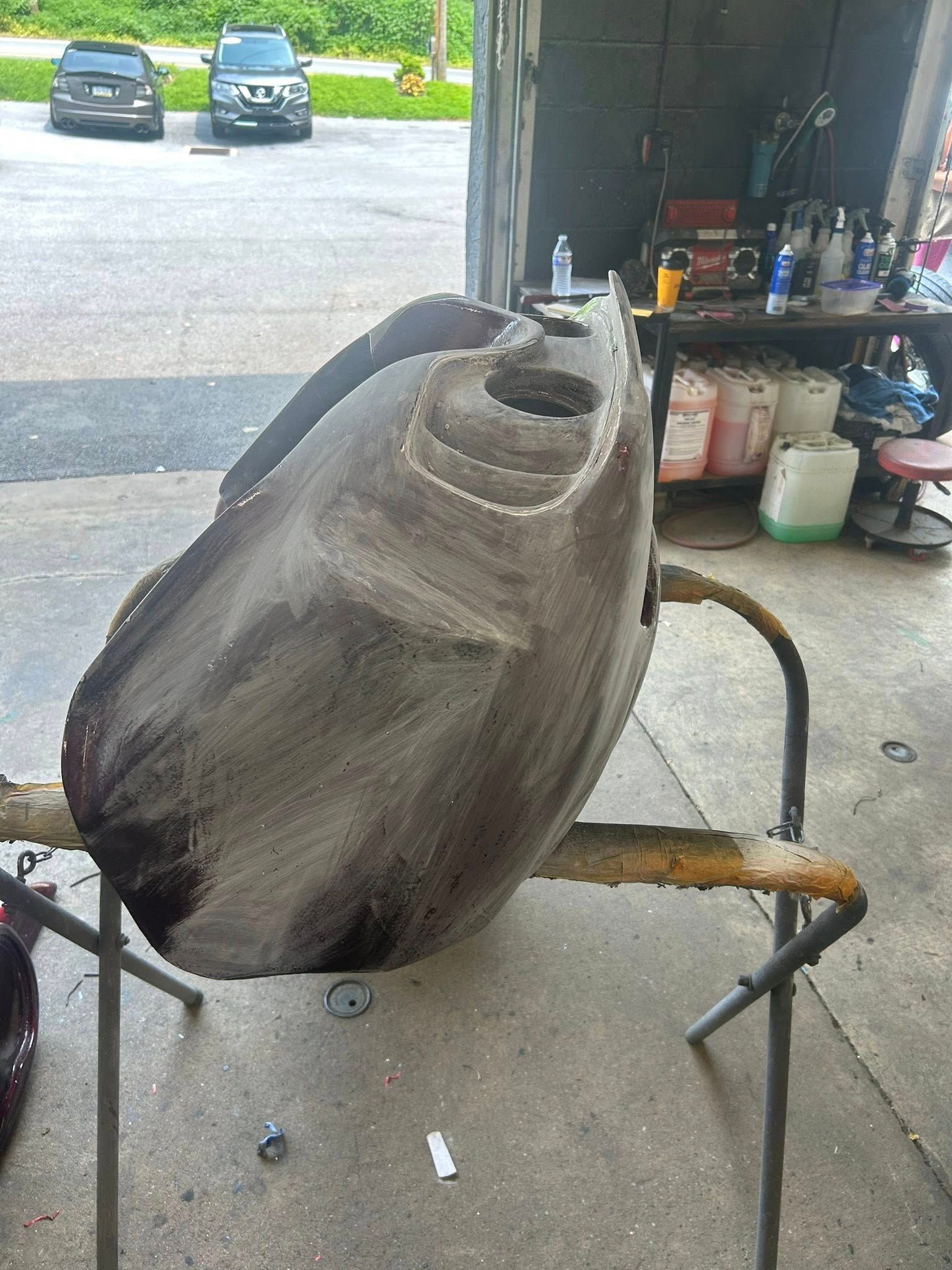 Large, gray vehicle part on a stand in a workshop, with cars and supplies visible in the background.