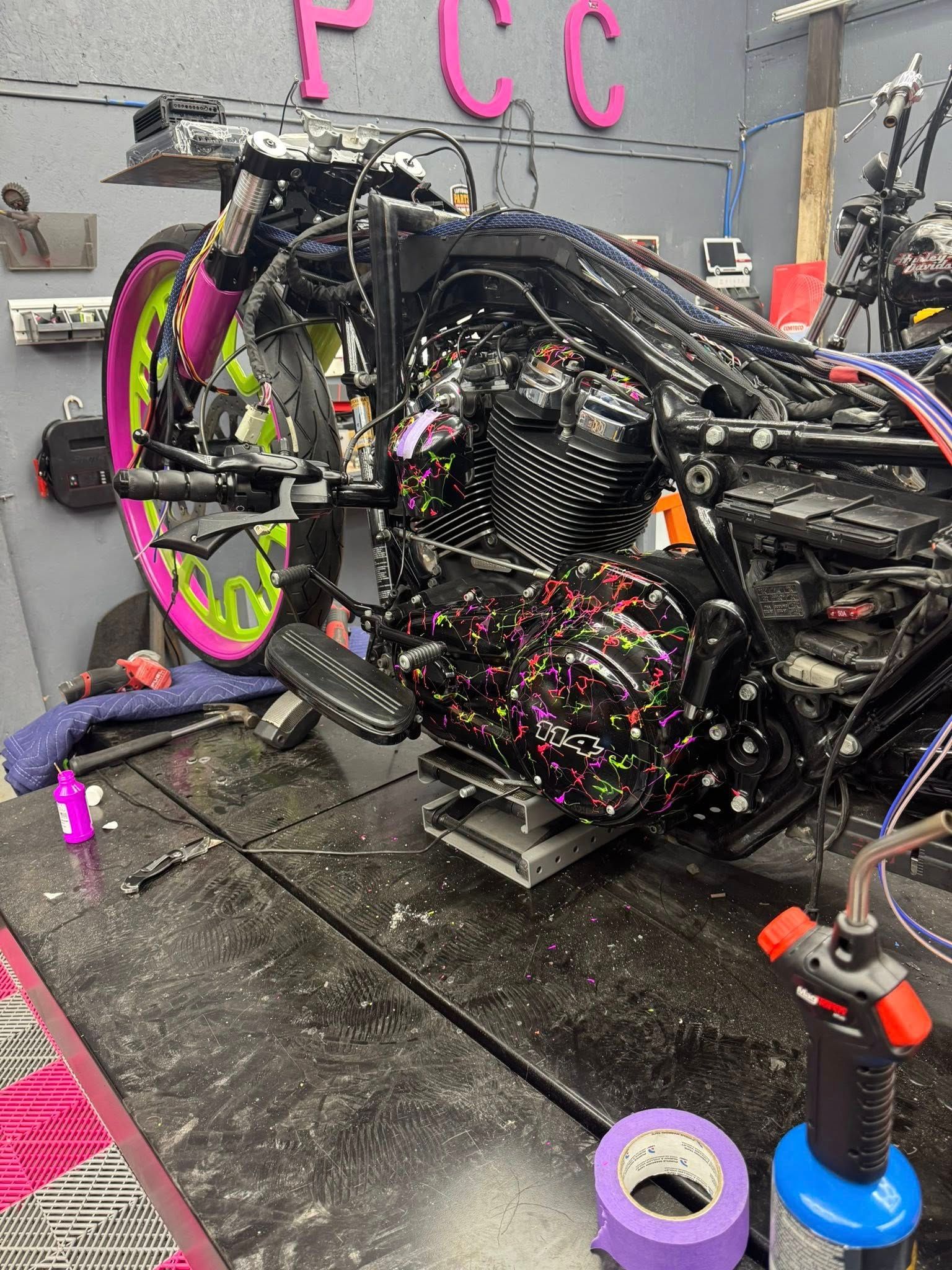 Motorcycle in shop, mostly disassembled, with pink and lime green wheels. Torch, tape, and tools are on the table.
