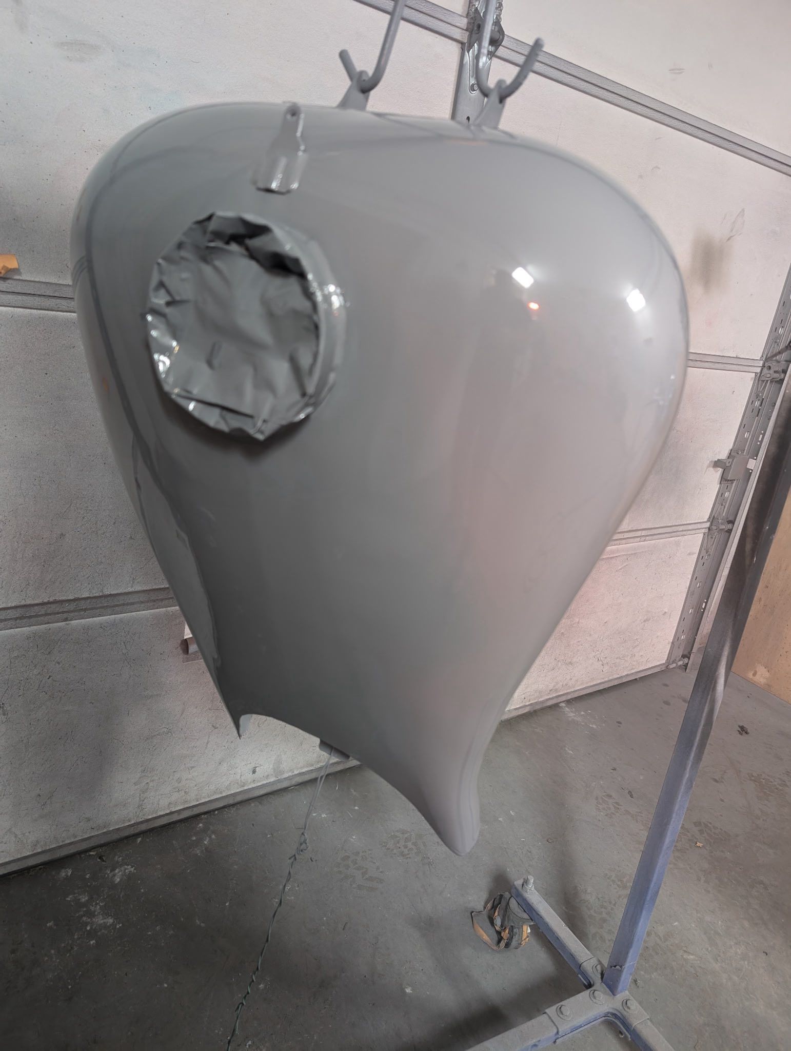 Gray motorcycle gas tank, freshly painted, hanging from a rack, with taped-over opening.