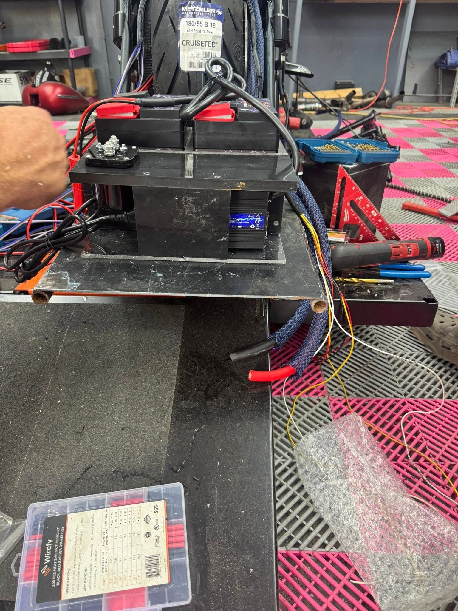 Motorcycle with batteries and wiring on a black platform, red and blue wires visible, workshop setting.