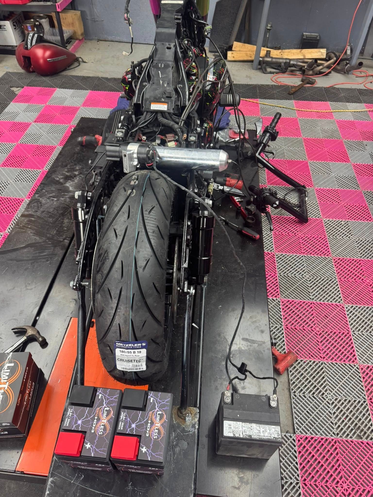 Motorcycle frame on a lift, rear wheel visible. Black frame with wires, battery, and boxes on the floor.