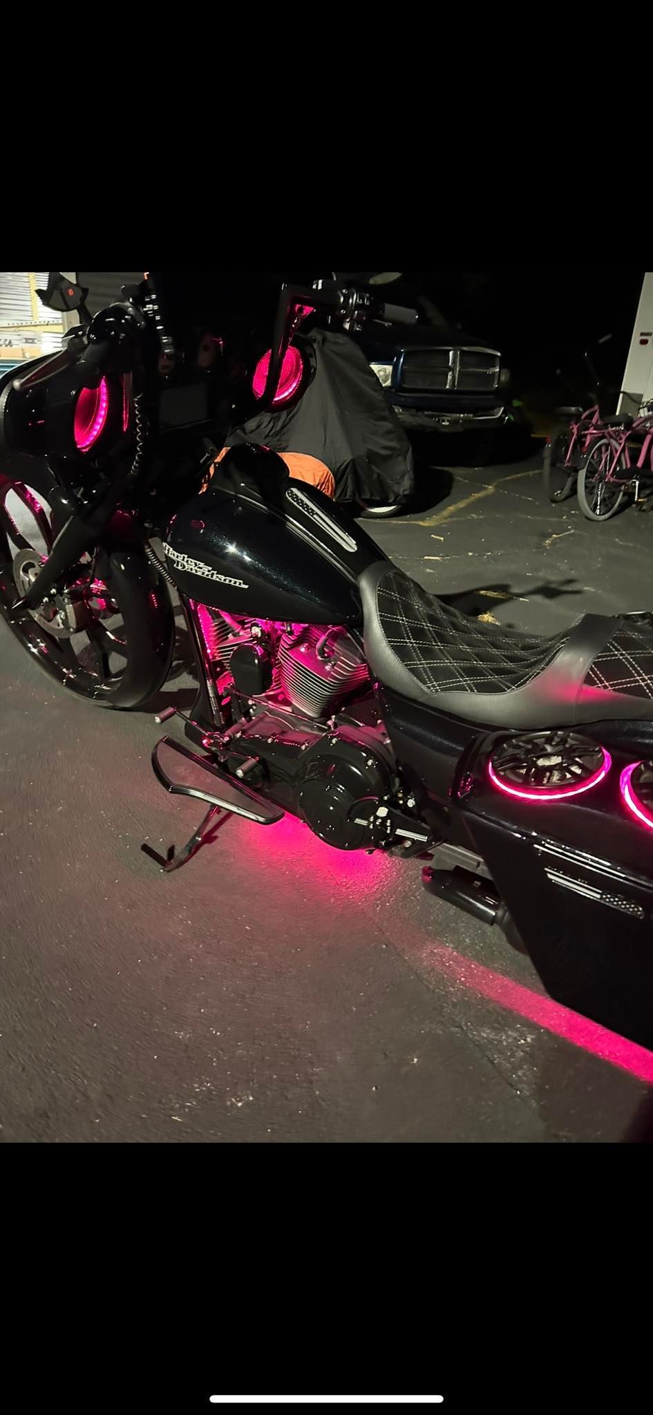 Customized motorcycle, black with pink neon lighting. Rider partially visible, parked outdoors at night.