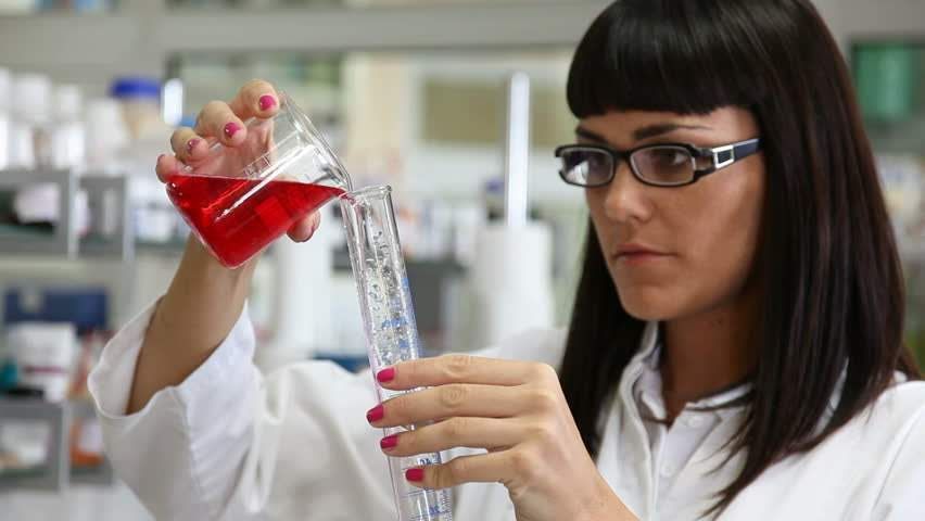 A woman in a lab coat is pouring a red liquid into a beaker.