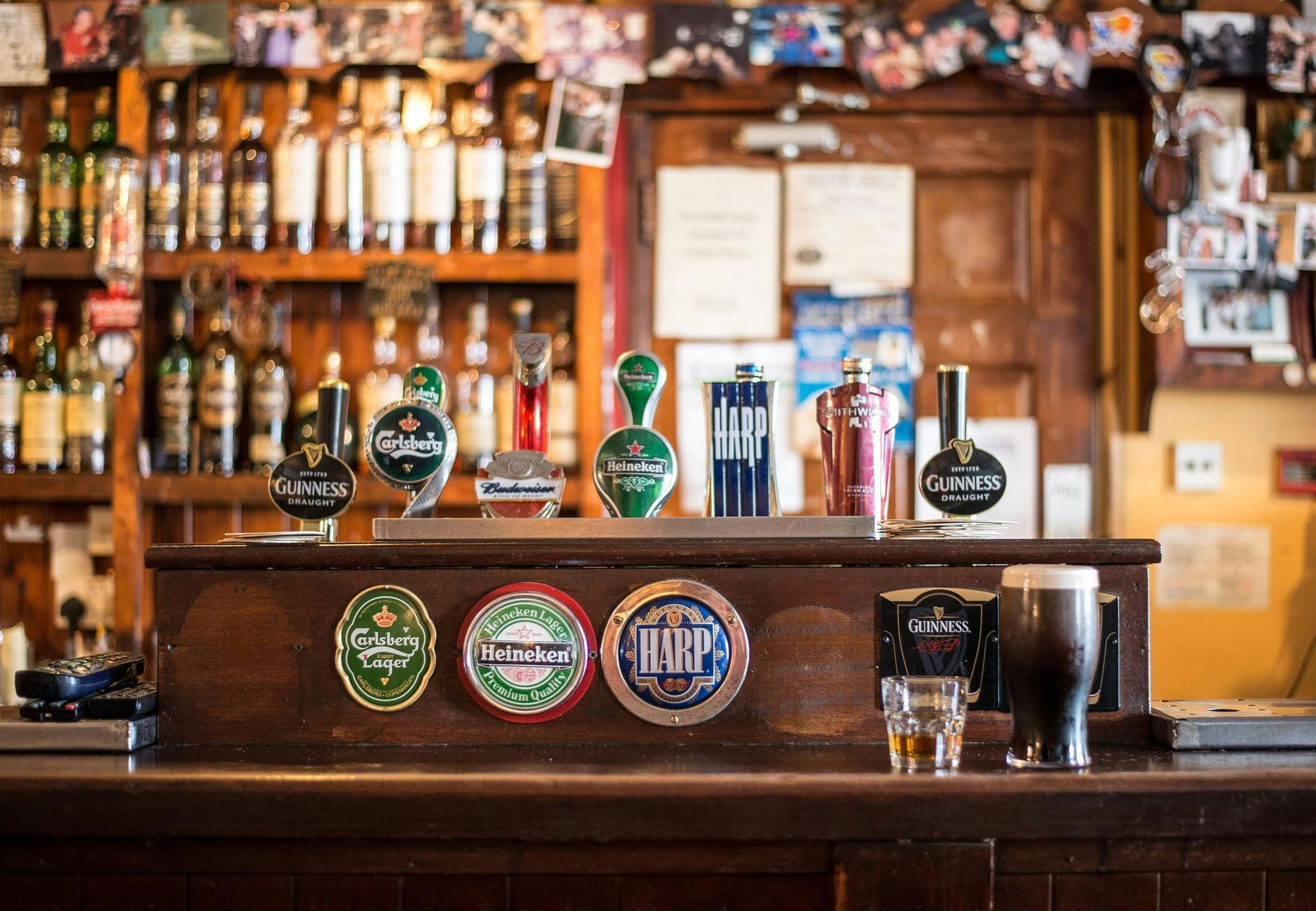A pub bar with beer taps, bottles, and a pint of Guinness. Wooden interior, warm lighting.