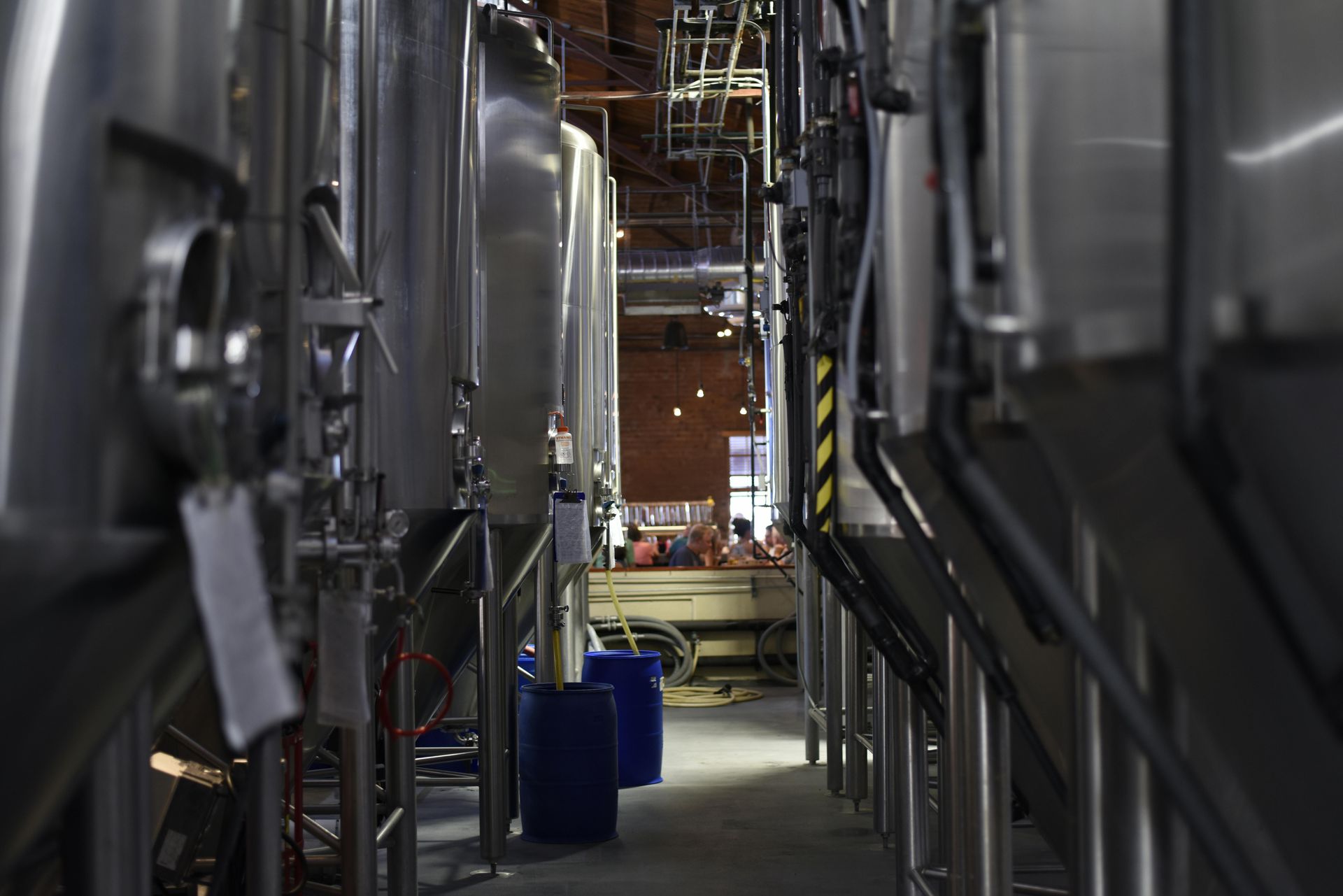 Inside a brewery, stainless steel tanks line a narrow walkway, with blue barrels and equipment.