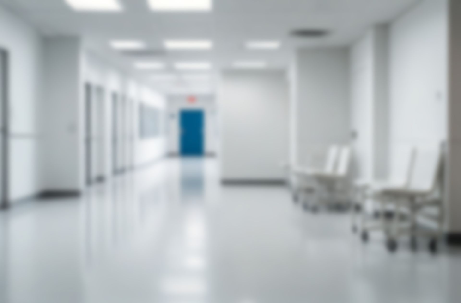 Blurred hospital hallway with white walls, doors, and floor, with blue door at the end.