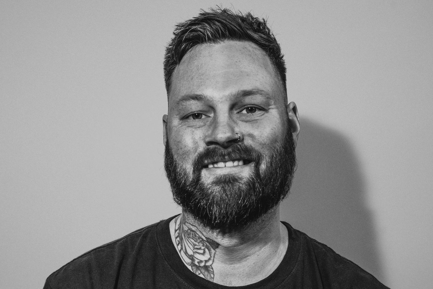 A man with a beard and a tattoo on his neck is smiling in a black and white photo.