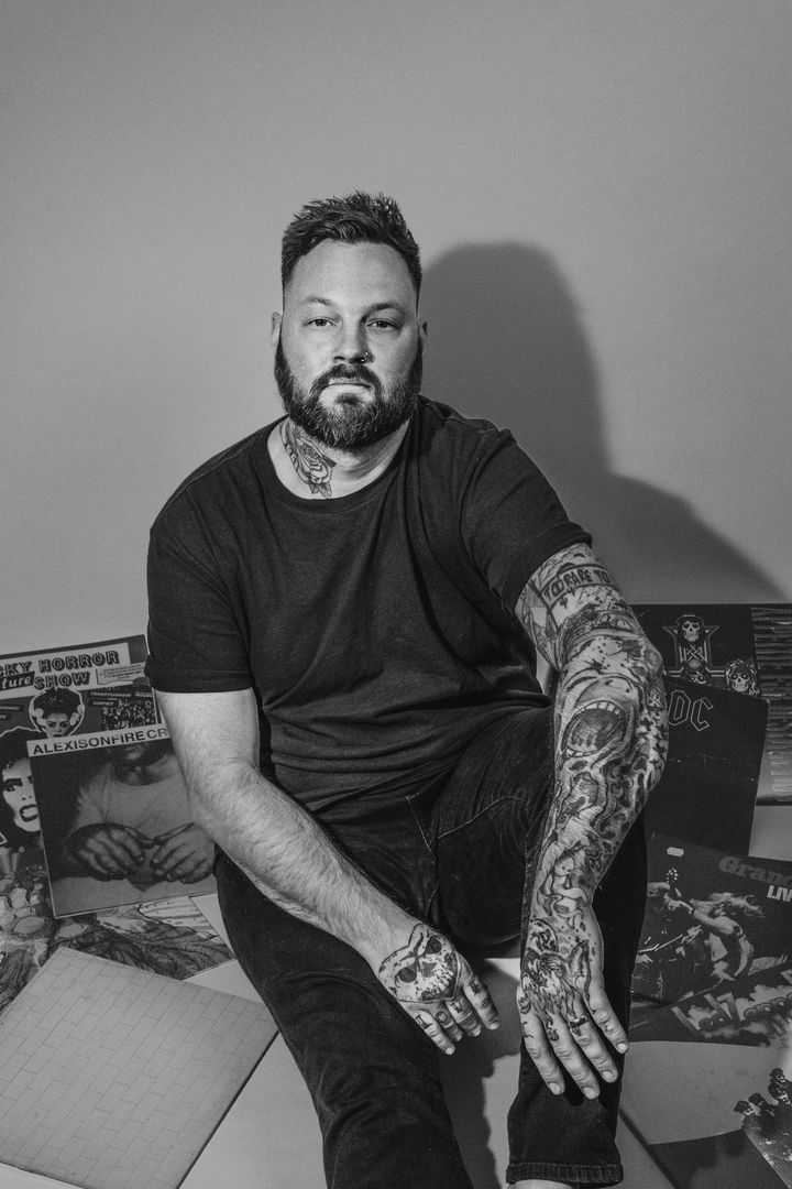 A man with a beard and tattoos is sitting on the floor in a black and white photo.