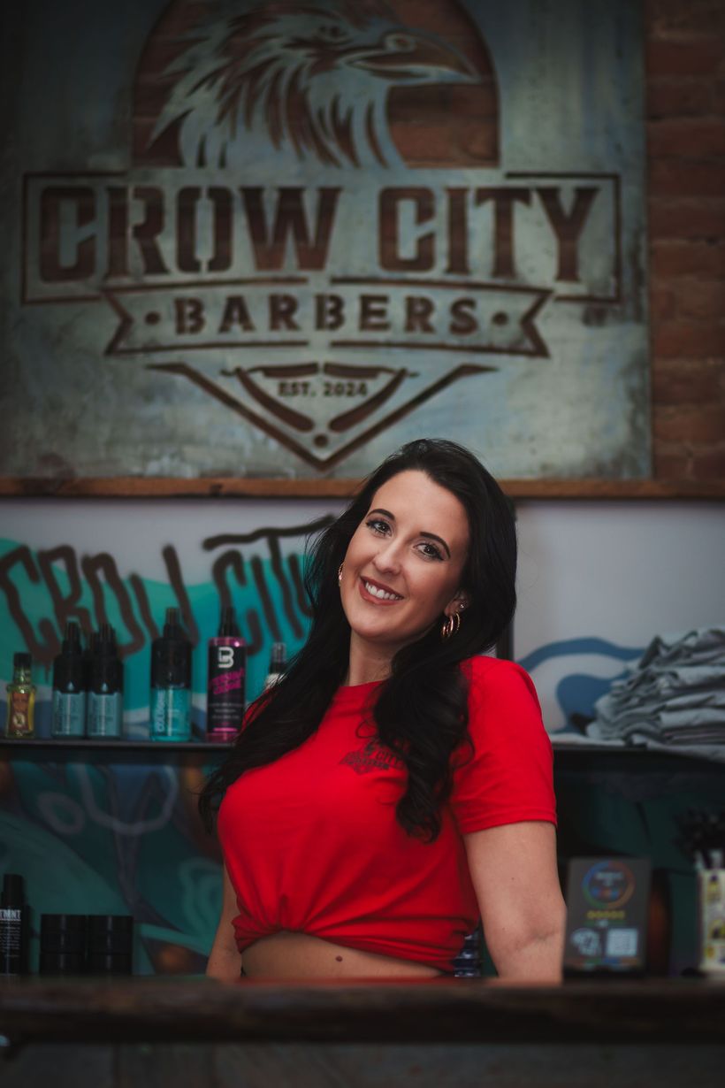 A woman in a red shirt is standing in front of a crow city barbers sign.
