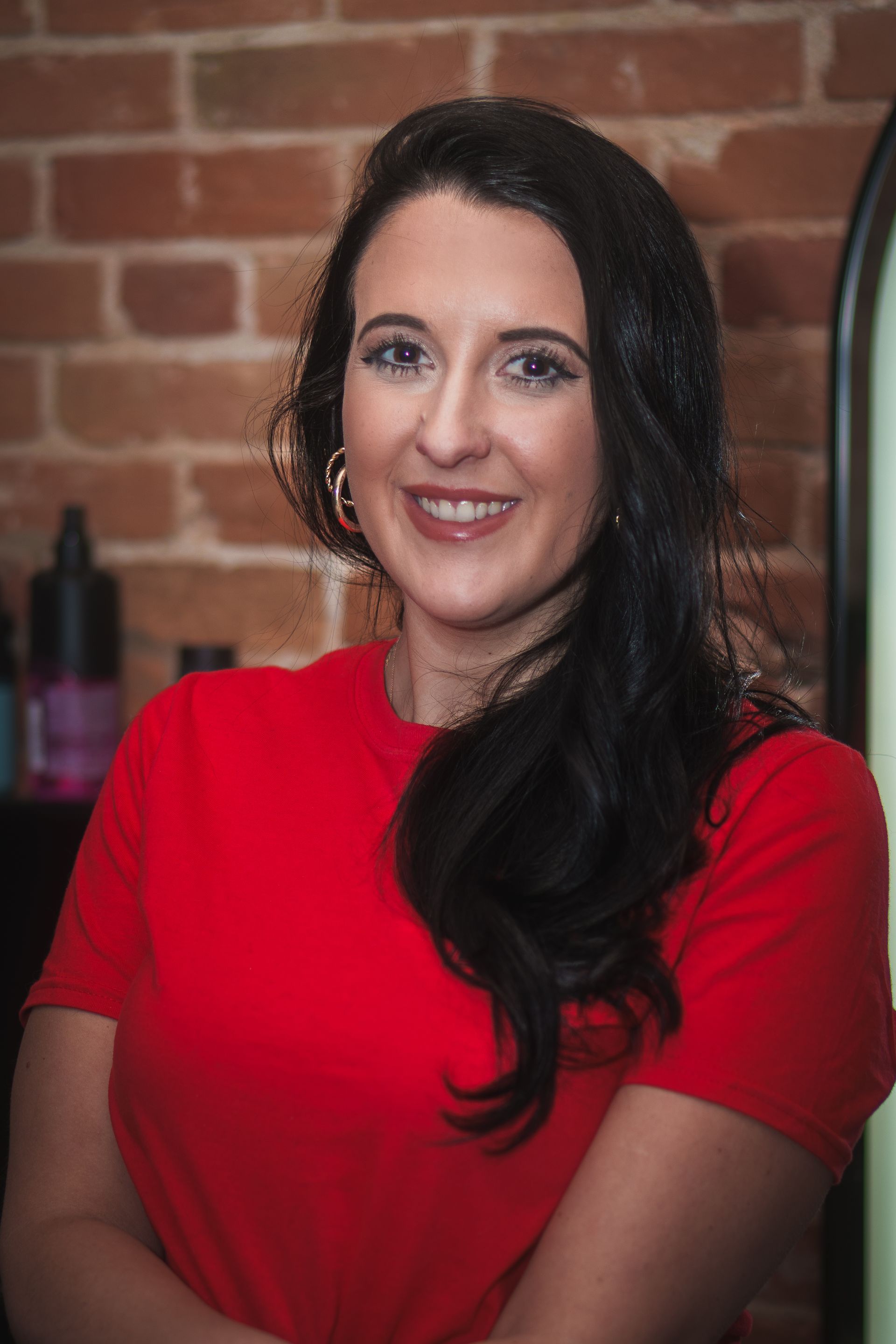 A woman in a red shirt is standing in front of a brick wall.
