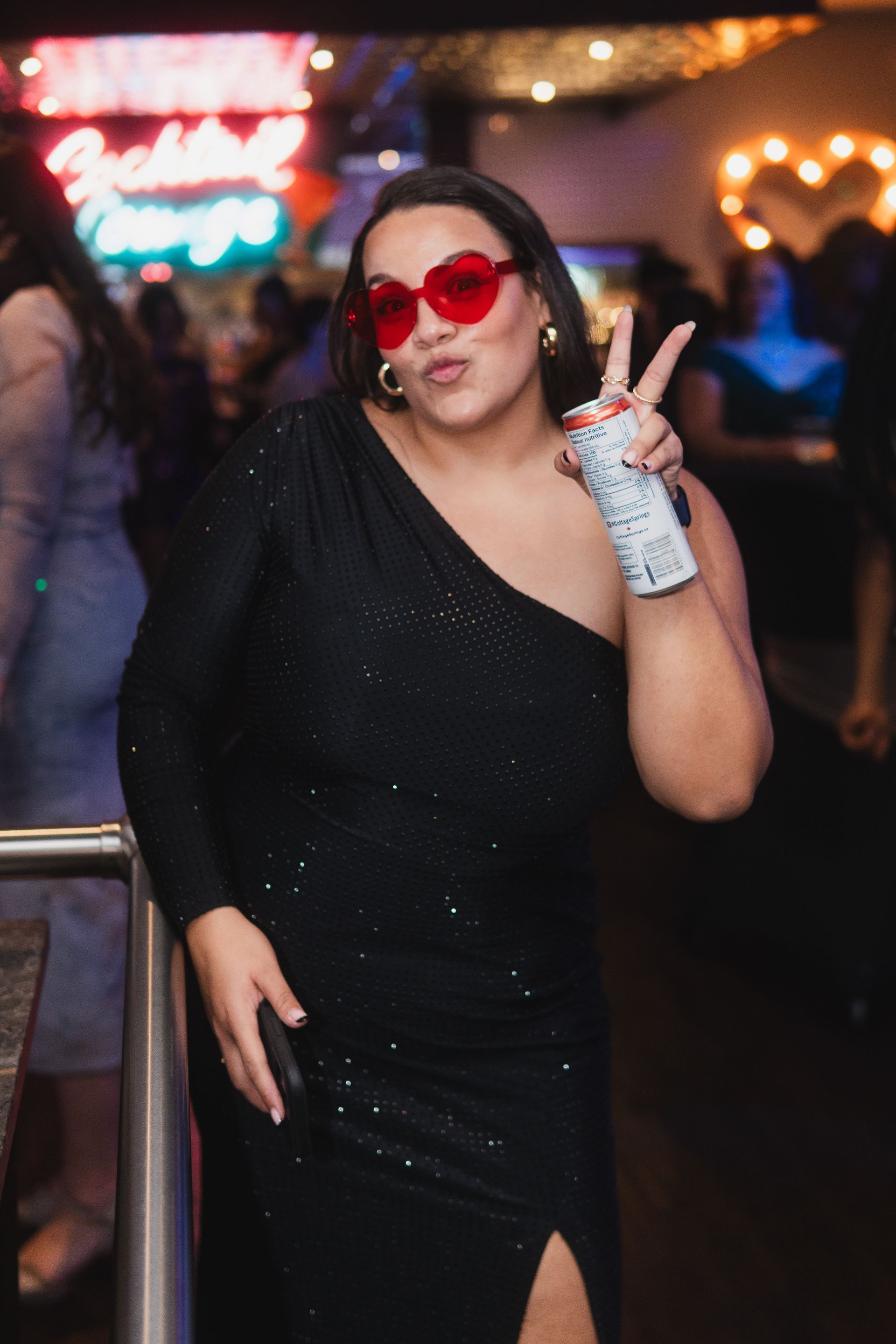 A woman in a black dress and red sunglasses is giving a peace sign.