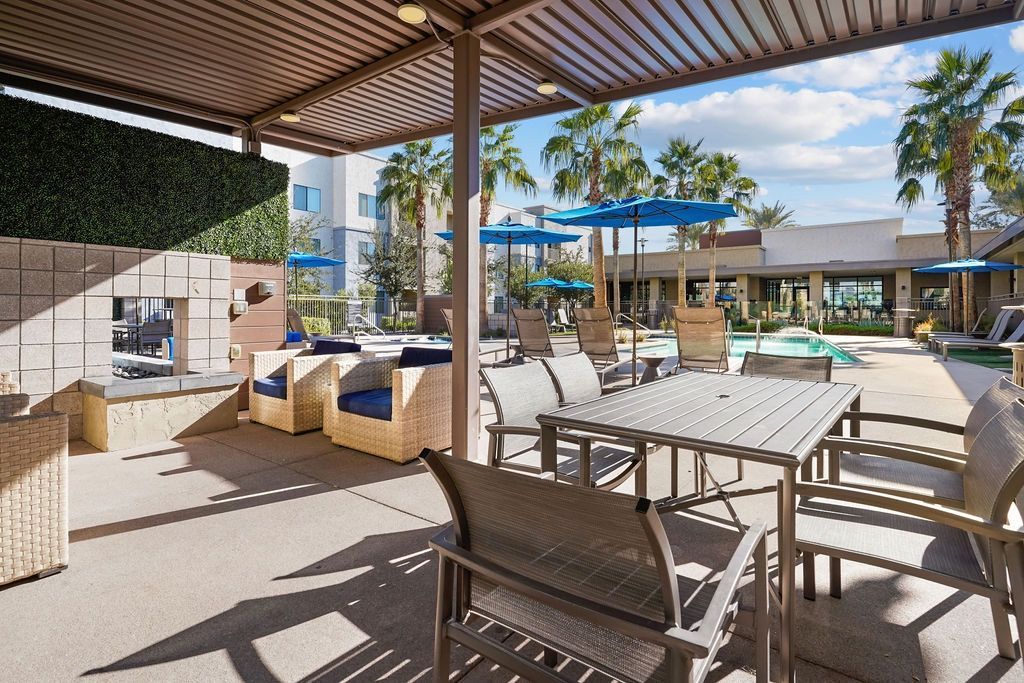 Outdoor community pool area with covered seating, lounge chairs, and blue umbrellas.