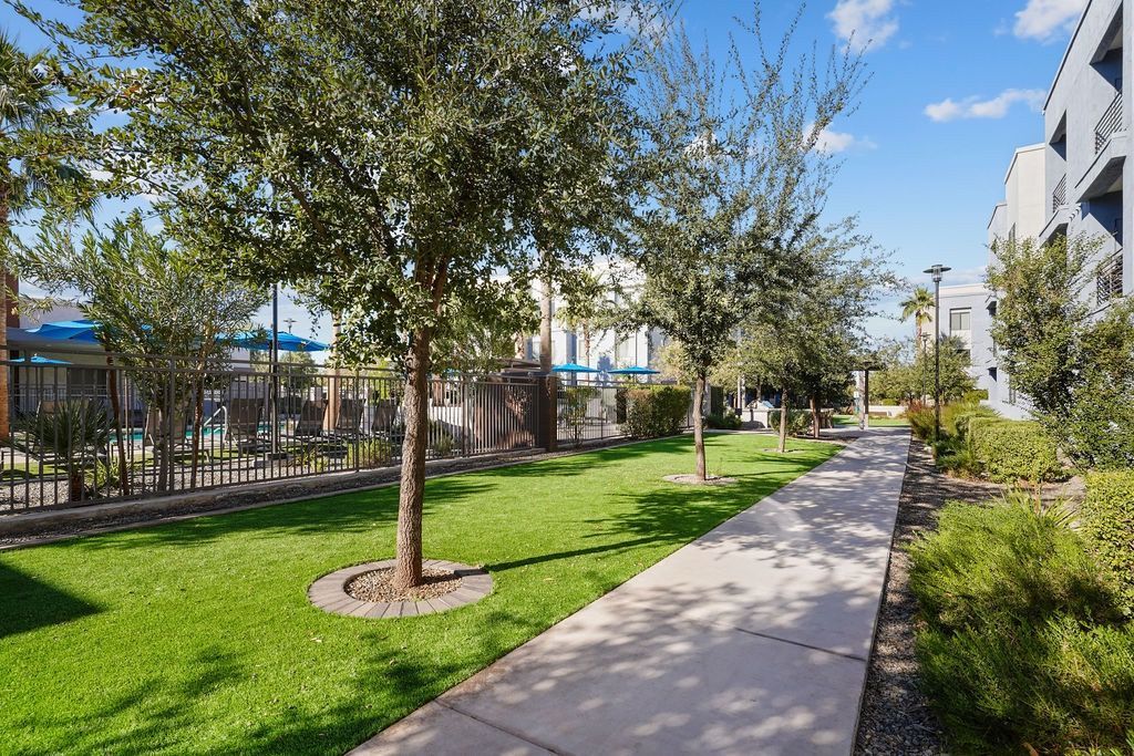 Pathway through a landscaped community courtyard with trees, grass, and a fenced pool area.