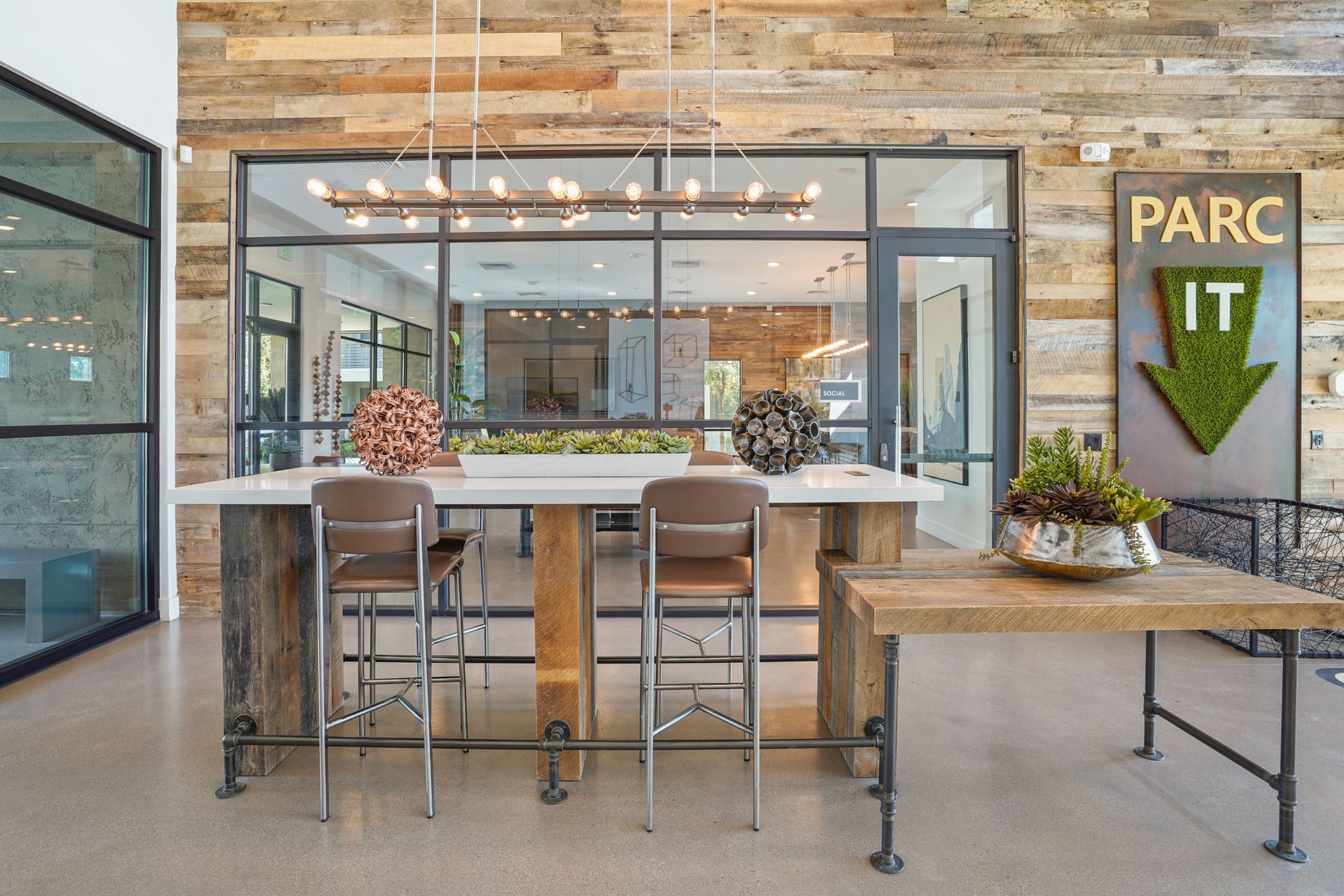 Reception area with a wooden feature wall, large windows, and a central table with bar stools at Parc South Mountain in Phoenix, AZ..